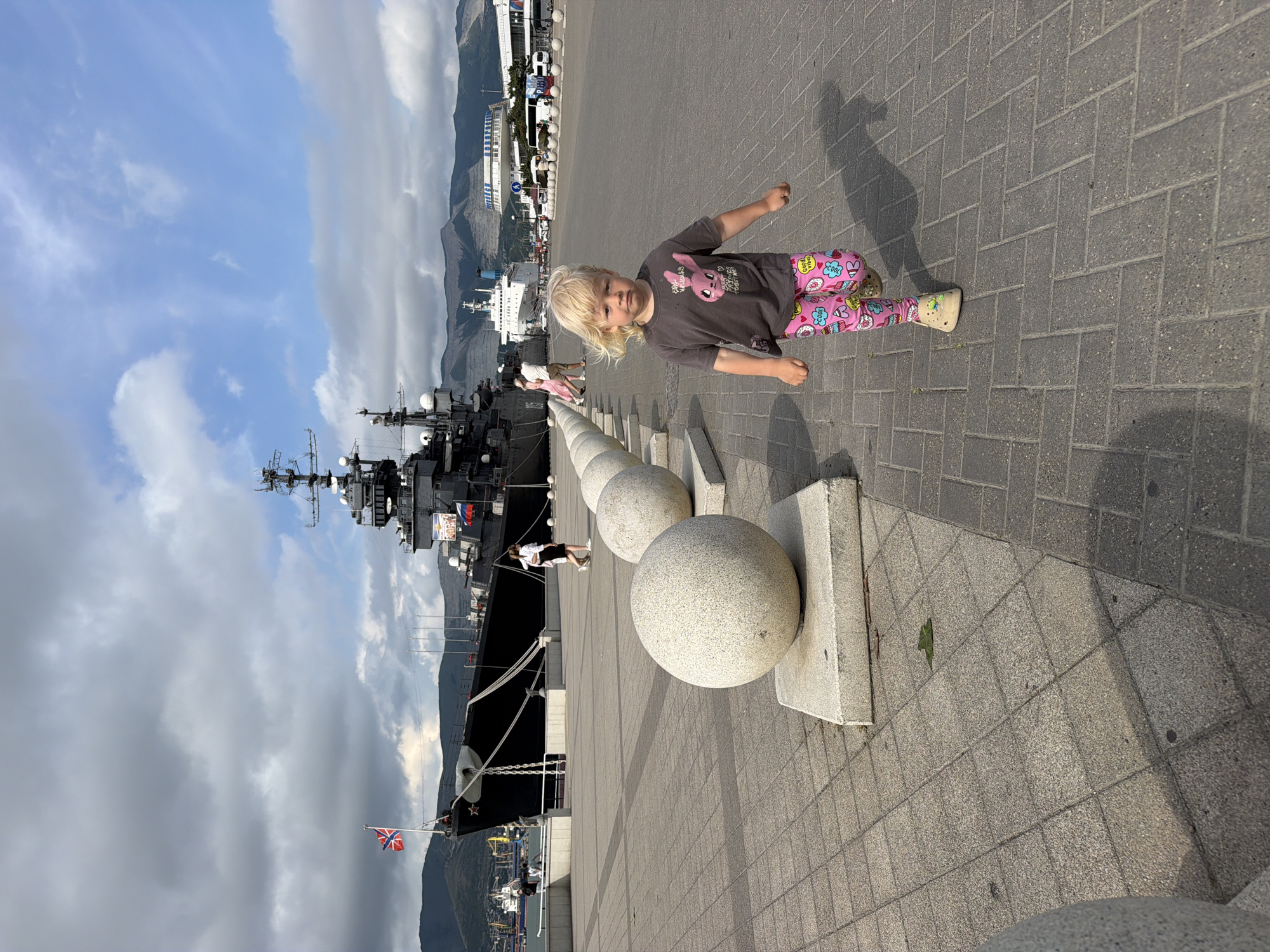In Novorossiysk, Russia, a young child with blonde hair runs towards the viewer on a paved walkway lined with large, round stone bollards. The child wears a dark gray t-shirt with a pink cartoon bunny graphic and pink leggings adorned with various colorful cartoon images, including cupcakes and the phrase "cool vibes." The child is also wearing beige clogs.
In the background, a large, dark-colored ship, possibly a museum ship, is docked. The ship has multiple decks and antennae, and the Russian flag is visible flying from its mast. Behind the ship, a white cruise liner is also docked, and mountains can be seen in the distance under a cloudy sky. The walkway is made of gray interlocking pavers, and the bollards are made of textured light gray stone, each resting on a white concrete base. Several other people are visible in the background, walking along the quay. The scene appears to be a public waterfront area.
