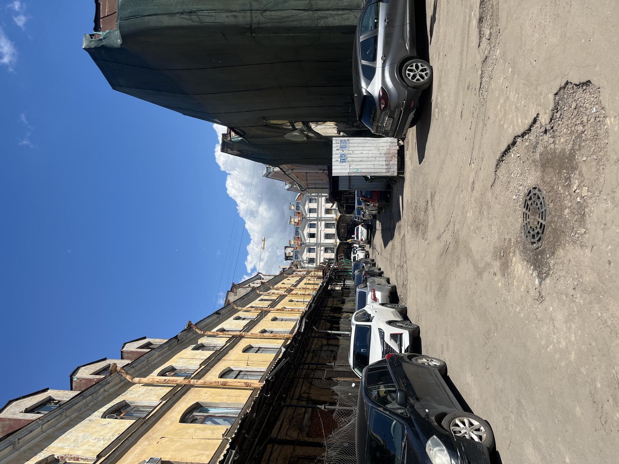 The image depicts a narrow street in Saint Petersburg, Russia, with an aged, yellow building on the left and a large structure covered in dark green netting on the right. The street is paved with asphalt and shows signs of disrepair, including potholes and a manhole cover. Several cars are parked along the street, including a black sedan, a white SUV, and a gray hatchback. The sky is blue with scattered white clouds. The architecture suggests an older, possibly industrial or residential area within the city. The overall impression is one of urban decay and limited vehicular access.