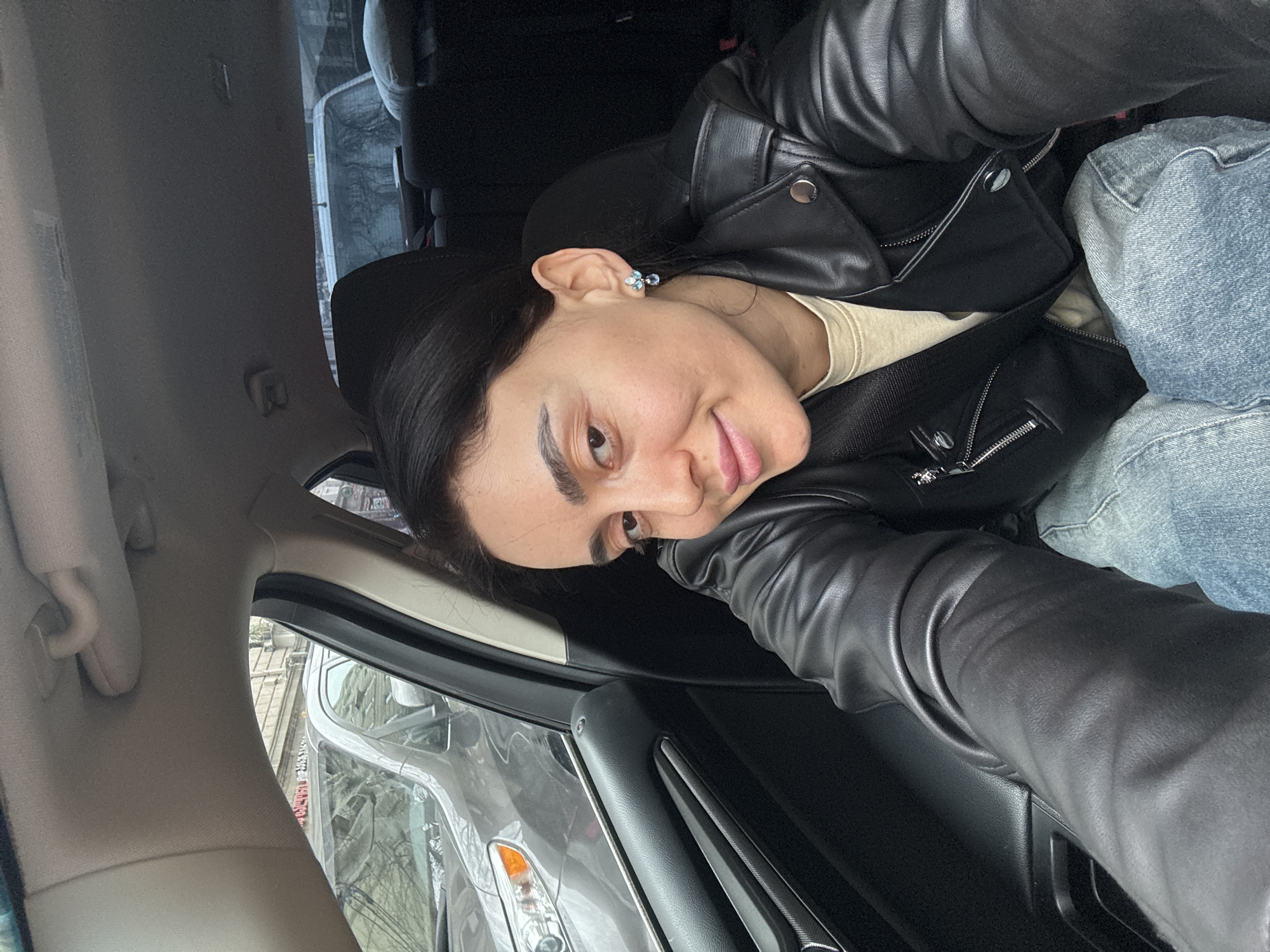 A female individual is situated in the front passenger seat of a vehicle, capturing a selfie. She has dark hair, light brown eyes, and a subtle smile, wearing a black leather jacket over a light beige top, light blue jeans, and light blue and silver earrings. The vehicle's interior is dark-toned with a light-colored headliner and visible back seats. Outside the front passenger window, a grey SUV is observed, reflecting bare tree branches. Beyond the vehicles, a multi-story building with older architectural details is visible. Faint reflections within the grey SUV's window reveal partial Georgian script on a red sign, confirming the location as a street in the city of Tbilisi, Georgia. The rear window shows another white vehicle and additional tree reflections. The scene is illuminated by diffused daylight, indicative of an overcast sky.
