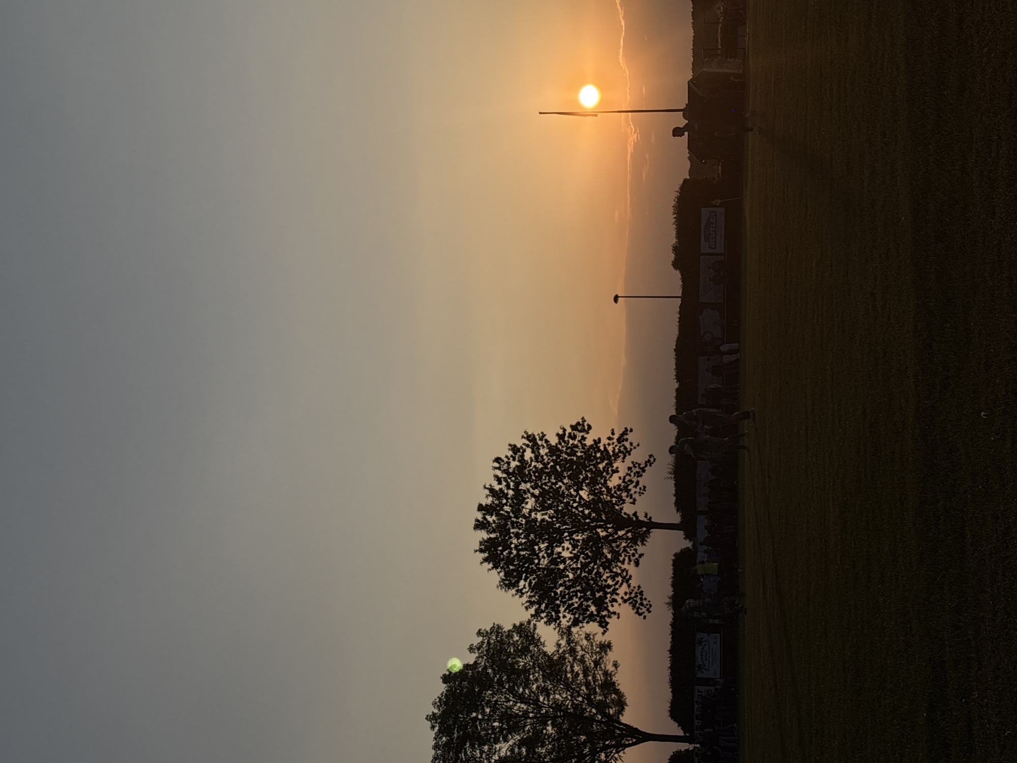 The image depicts a sunset scene at a location in Wegberg, Germany. A partially obscured sun shines above what appears to be a sports field, marked by a flag pole. Along the edge of the field are trees. In the background is a perimeter fence with advertisements. Silhouetted figures of individuals are observable on the field. The sky gradient ranges from grey to orange.
