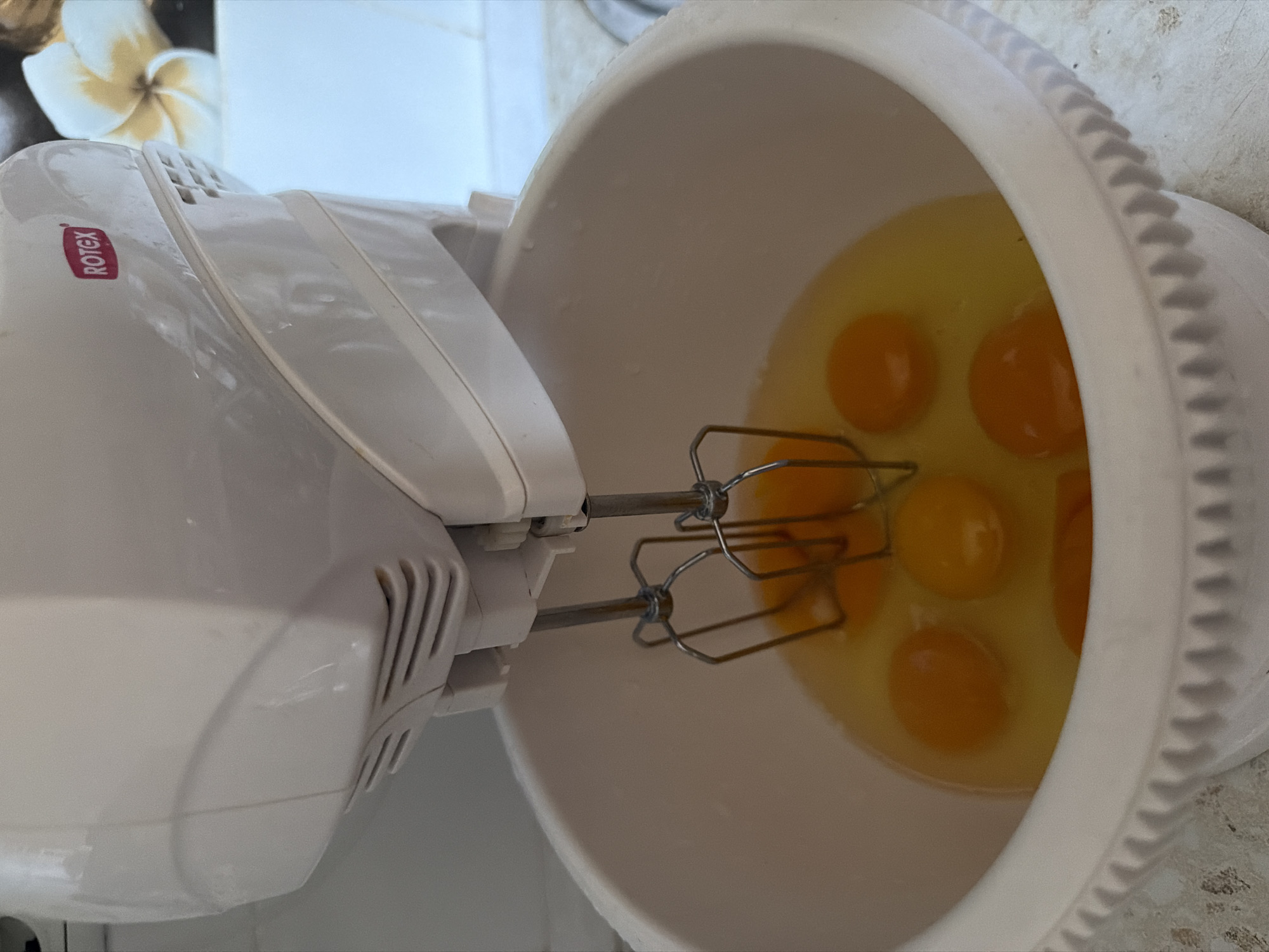 An overhead, close-up shot shows a white hand mixer with beaters submerged in a white mixing bowl filled with raw eggs. The mixer has a red "Rotex" logo on its side. The bowl is mostly filled with egg yolks and whites, with at least five yolks visible. The mixer is positioned to the left of the bowl, with its beaters in the center. The background is blurred, showing a light-colored surface and a white and yellow flower. The scene appears to be in a kitchen, as suggested by the countertop and the presence of a mixing bowl and mixer. There are no people visible in the image. The lighting suggests it might be daytime.