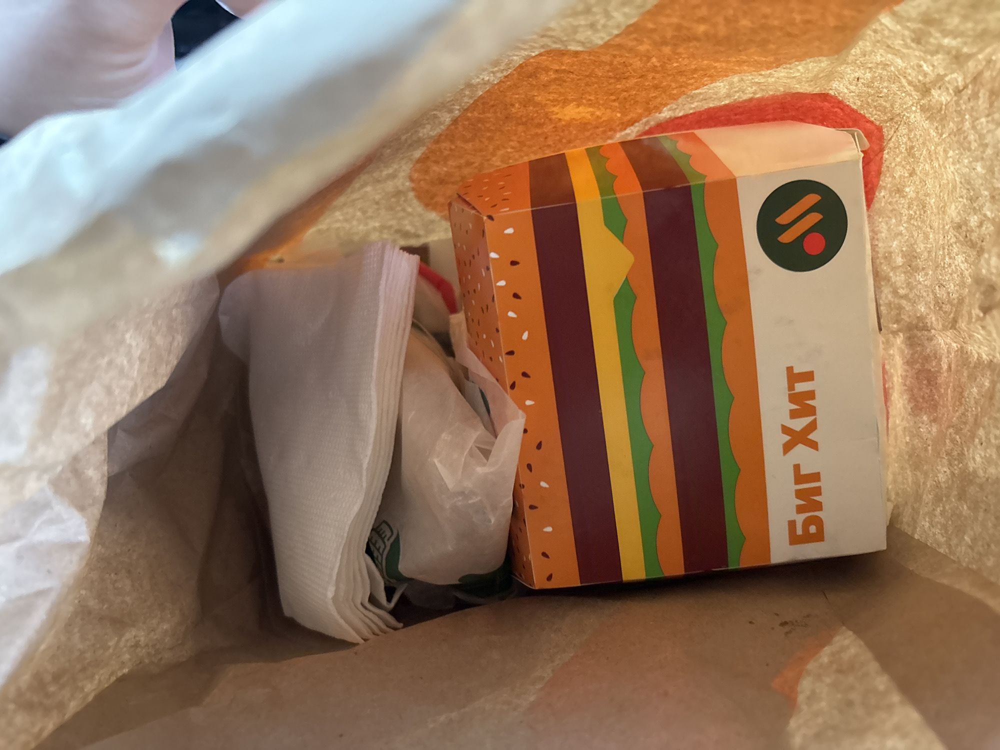 Inside a brown paper bag, a box labeled "Биг Хит" (Big Hit) with a stylized burger graphic sits next to a stack of white napkins and a small plastic package. The packaging design suggests the food is from a fast-food restaurant. While the image context hints at the city of Rusanovo, Russia, this cannot be definitively confirmed from the image alone.