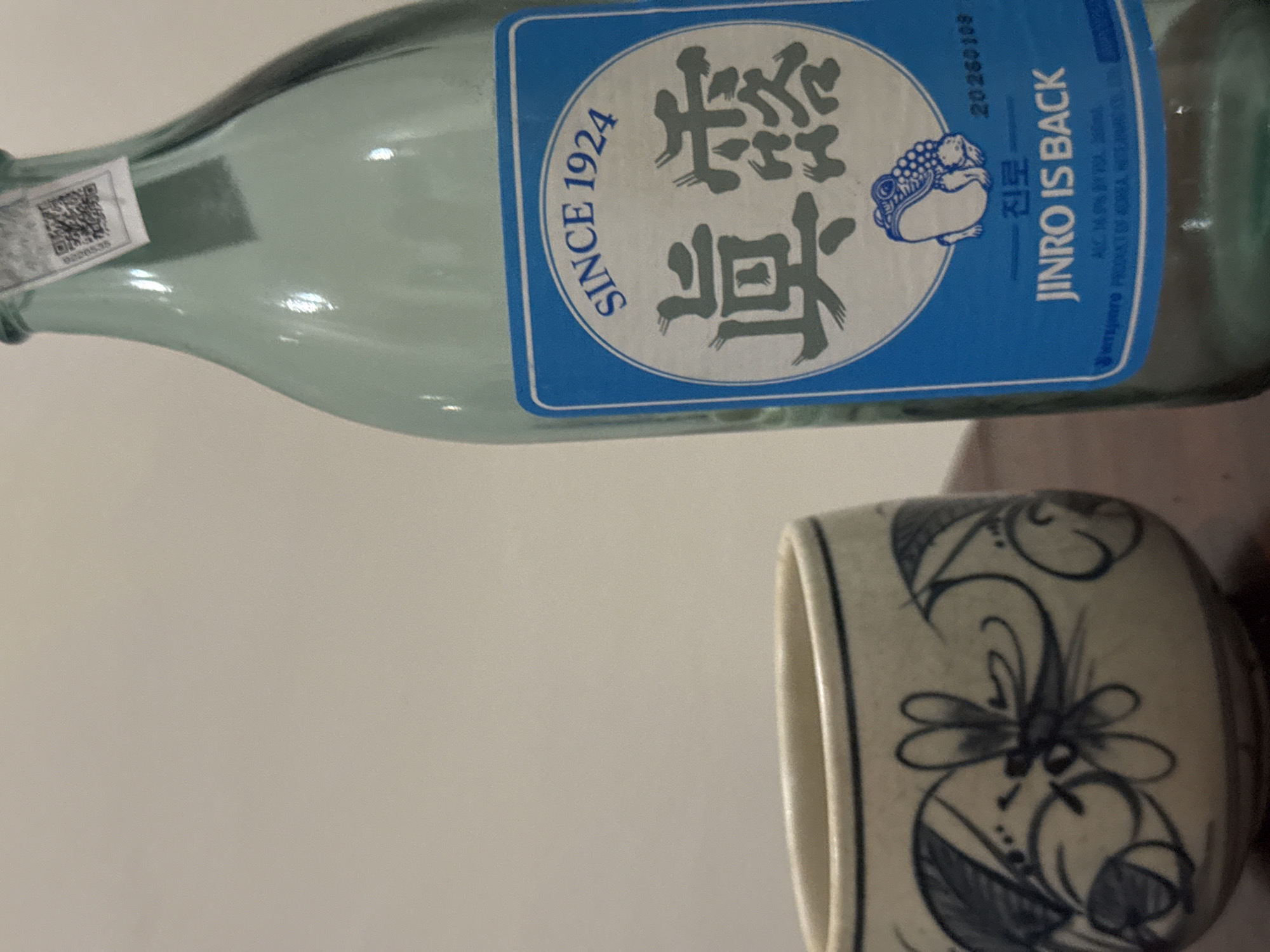 The image shows a bottle of Jinro soju and a ceramic cup. The bottle has a blue label with the words "SINCE 1924" and "JINRO IS BACK". The cup has a floral pattern. The background is a plain, light color. The location is not specified in the image.