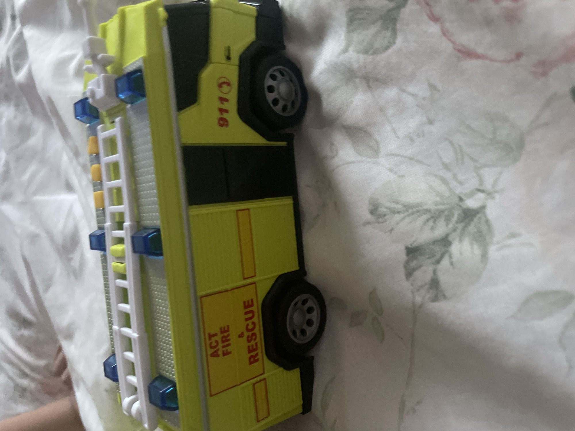 A bright yellow toy fire truck with blue lights on top and the text "ACT FIRE & RESCUE" on its side sits on a white and floral patterned surface. The truck has a ladder on top and black wheels. The background is out of focus, but appears to be a bed with white and pink floral bedding.