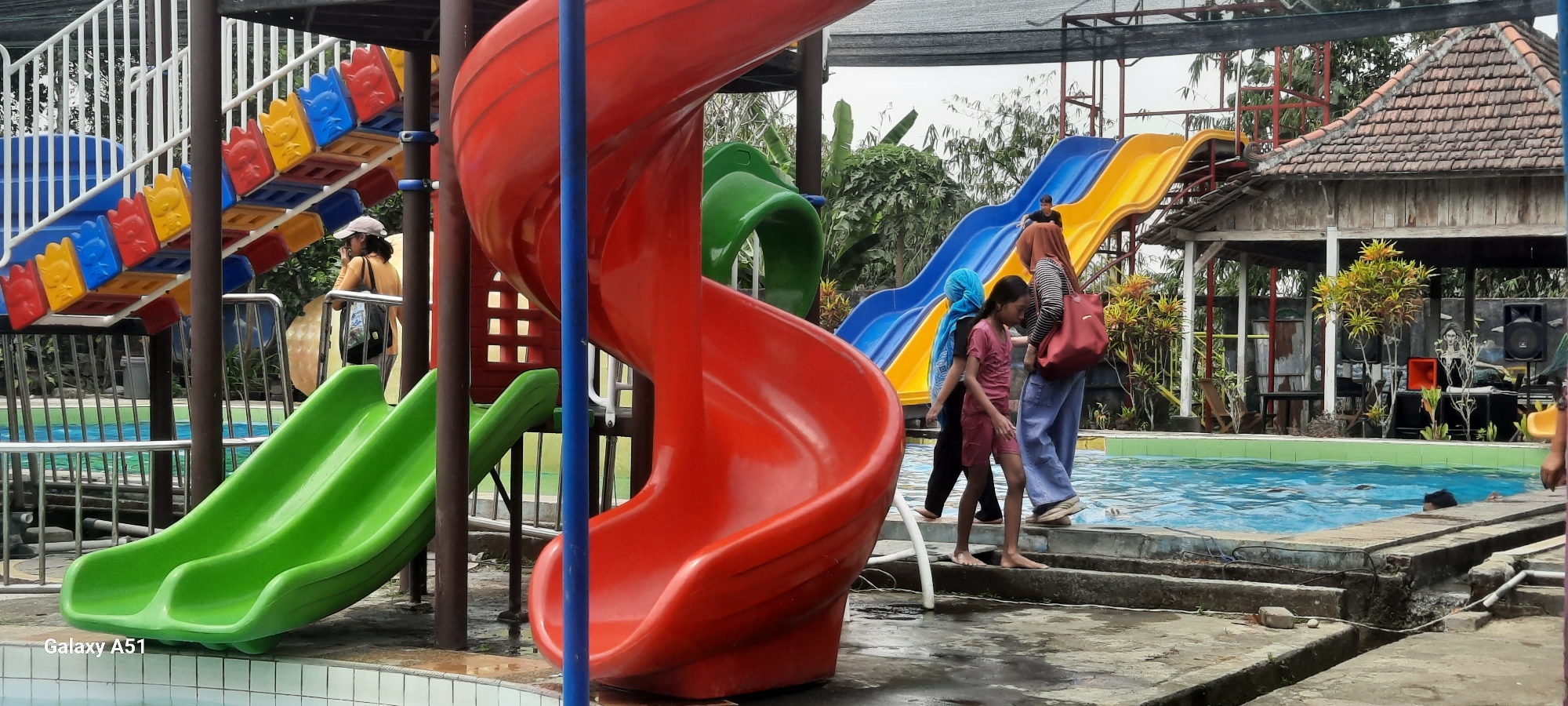 The image depicts a vibrant water park in Popoh, Indonesia, featuring a multi-level playground with colorful slides. In the foreground, a bright red spiral slide dominates the left side of the frame, with a green double slide to its left. To the right, a blue and yellow slide descends towards a swimming pool. Several people are present. In the mid-ground, a young girl in a pink shirt and shorts, accompanied by two adults wearing hijabs and casual attire, walks near the edge of the pool. Another individual in a black shirt is seen sliding down the blue and yellow slide. Further in the background, a woman in a light-colored cap and orange shirt stands near the stairs of the playground structure. The environment is lush with green trees and vegetation surrounding the pool area. A rustic-looking wooden structure with a tiled roof, possibly a pavilion or changing room, is visible on the right side, with sound equipment and speakers set up in front of it. The ground around the pool is wet and appears to be concrete. The overall scene suggests a recreational day at the water park.