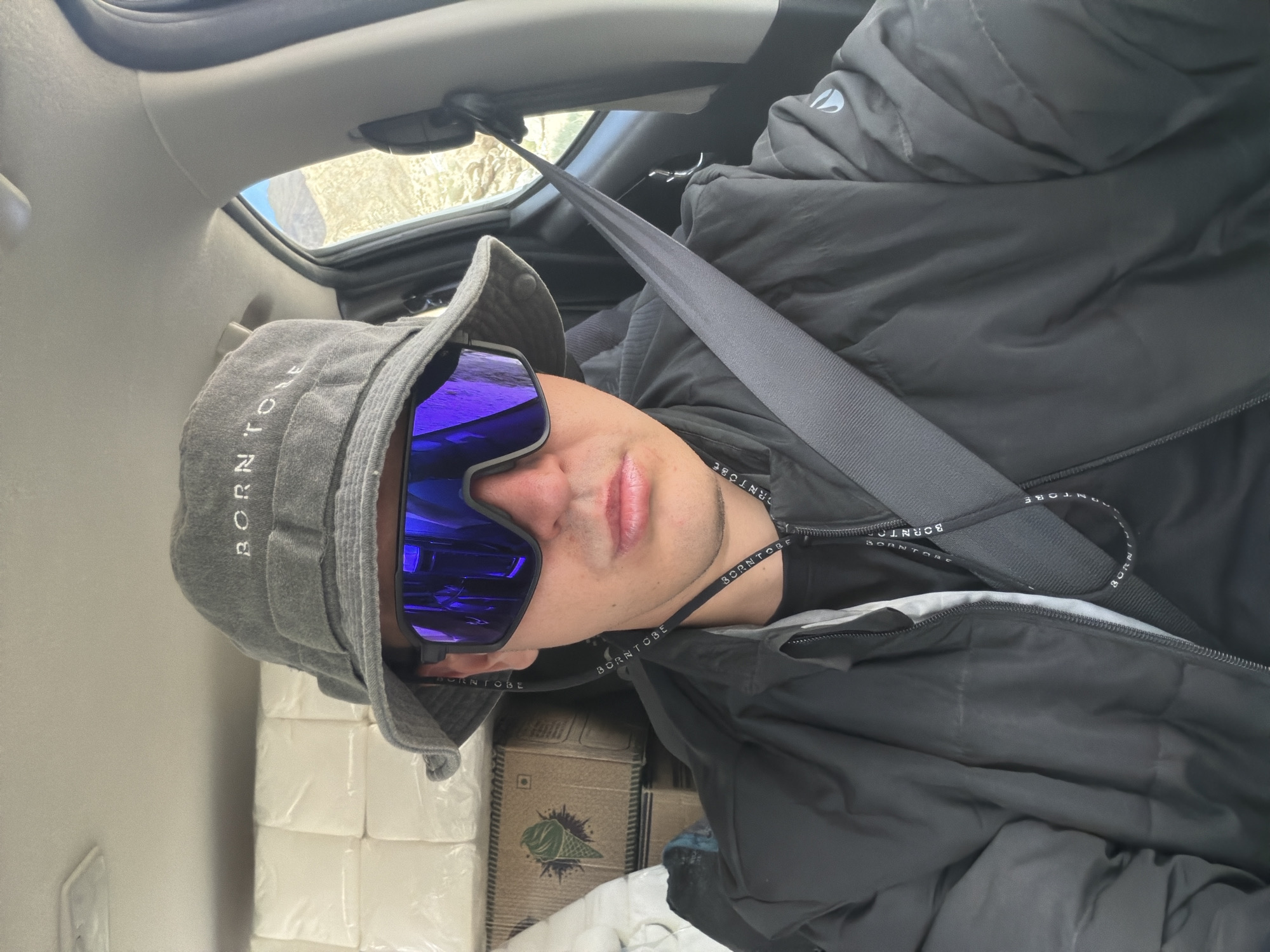 A person wearing a grey bucket hat with "BORN TO BE" embroidered on it, mirrored sunglasses with blue lenses, and a black puffy jacket is seated in the driver's seat of a vehicle. The vehicle's interior is visible, with stacks of white packaged goods and cardboard boxes in the background. A grey seatbelt is fastened across the person's chest. The environment outside the vehicle, visible through the open window, appears to be a dry, mountainous region. The person's mouth is slightly open, and they are looking directly at the camera.