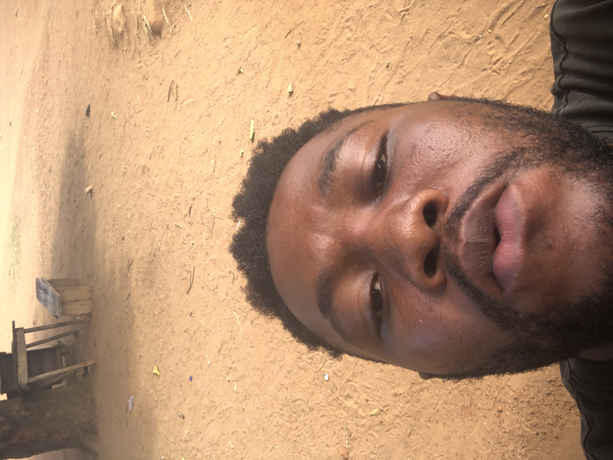 A man's face is prominently featured in the image, positioned against a backdrop of sandy or dirt ground. A rudimentary wooden structure is partially visible to the left of the frame. The location of the city is Unknown, Unknown.
