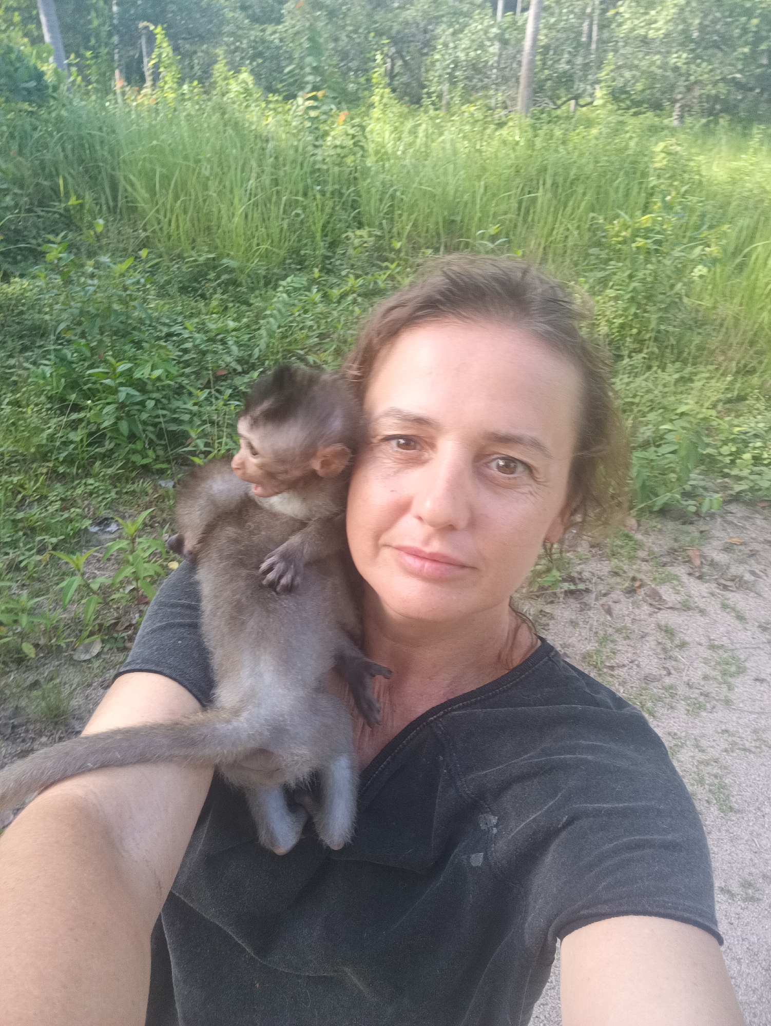 A woman holds a small monkey on her shoulder. The woman has fair skin, light brown hair, and is wearing a black v-neck t-shirt. The monkey is gray-brown, with a long tail that drapes down the woman's arm. The background is a mix of green foliage and a sandy path. The location is likely in Baan Thong Nang, Thailand.
