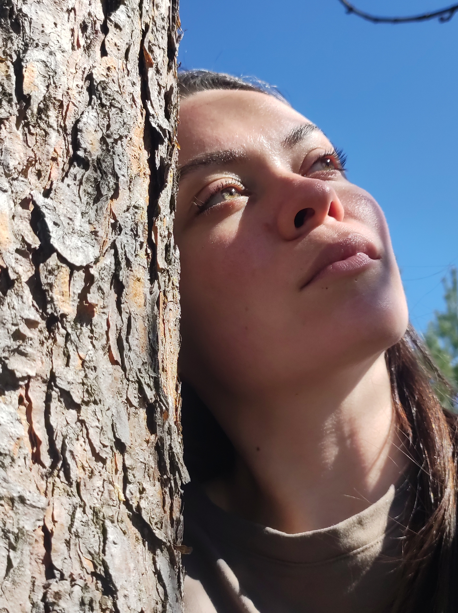 A person with long brown hair is leaning against a tree on a sunny day. The person is looking up and has a piercing in their ear. The tree has rough bark. The sky is blue with some visible branches. The location cannot be determined.