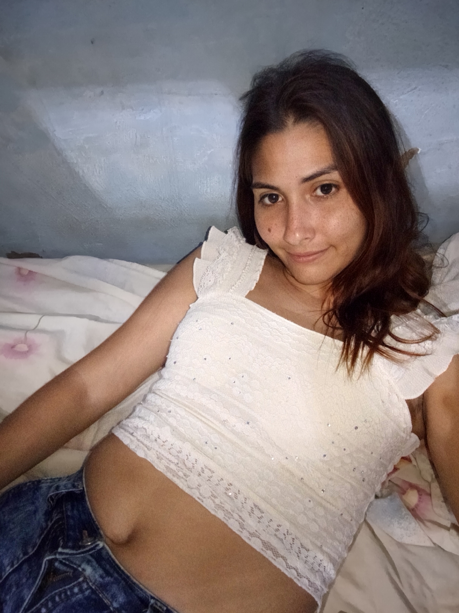 A young woman with brown hair and brown eyes is lying on a bed, looking at the camera. She is wearing a white lace top and blue jeans. The room is dimly lit, and the background is a textured blue wall. There are some flowers on the bedding.