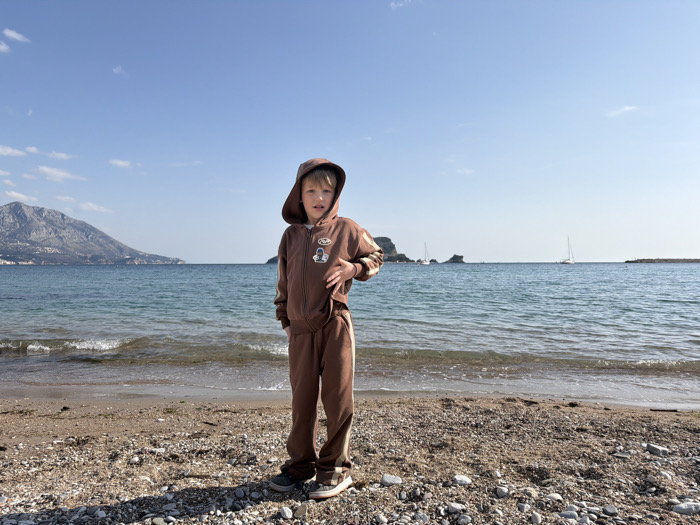 The scene is a sunlit gravel and sand beach, identified as Slovenska Plaža in Budva, Montenegro. A light-skinned child, approximately 5-8 years old, stands centered, facing the viewer. The child wears a brown hooded tracksuit with the hood up; their left hand is positioned near their chest, and their mouth is slightly open. The immediate background features the Adriatic Sea with gentle waves reaching the shore. In the mid-distance, several rocky islets and two white sailboats are visible on the water. A large mountain range dominates the far left background, while a distant headland or breakwater is present on the far right. The sky is clear blue with minimal cloud presence.