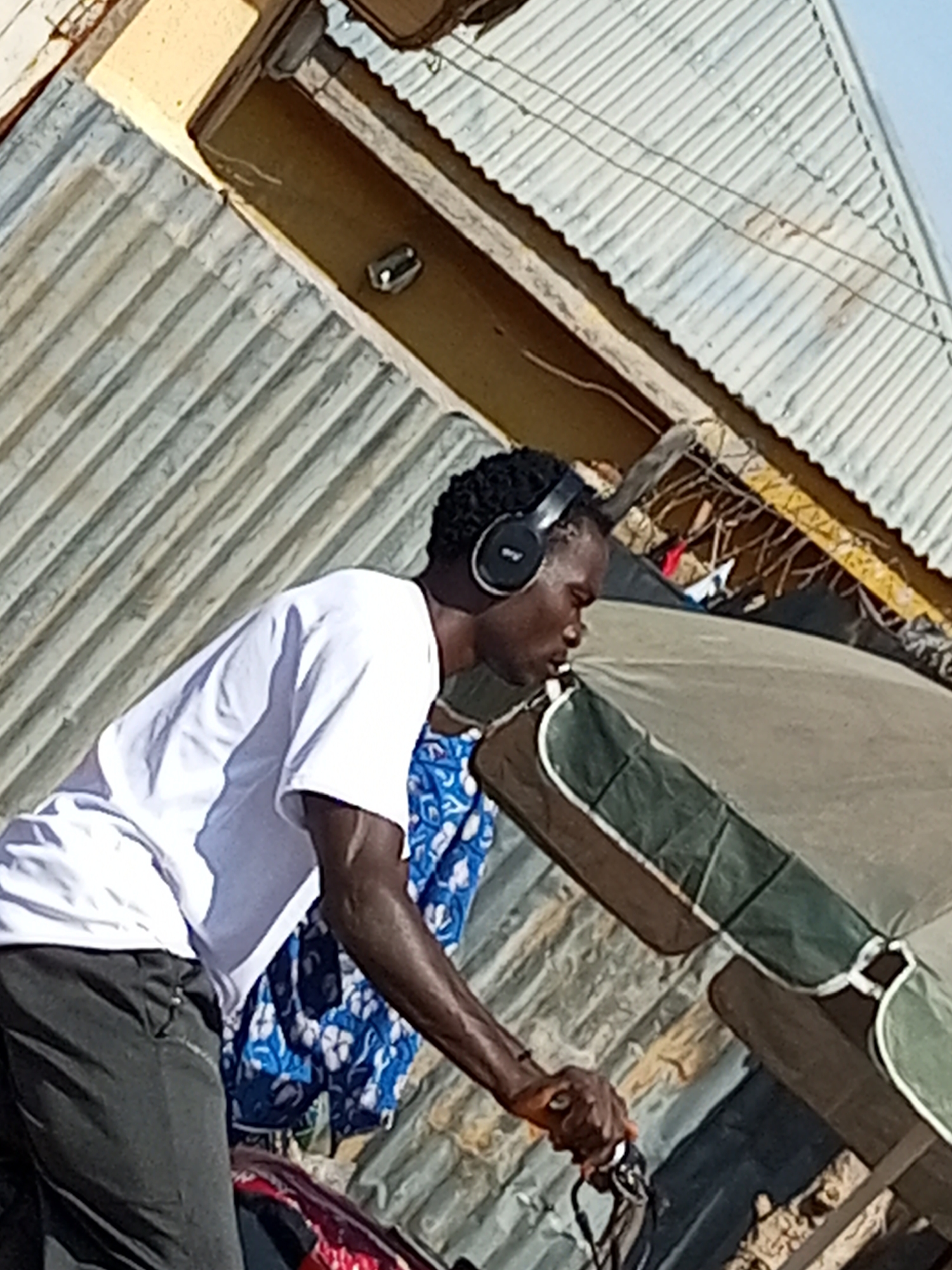 A young Black man wearing headphones and a white t-shirt is captured in a medium shot from the side, looking downwards as if engaged in a task or listening intently. His dark hair is styled in short, curly dreadlocks. He is wearing dark gray pants. His left arm is extended, and his hand is holding onto a dark object, possibly part of a bicycle or motorcycle handlebar, with cables attached.
The man is situated outdoors, in what appears to be a somewhat informal urban environment. Behind him, there is a structure with corrugated metal roofing, some of which is discolored and weathered. A large, light-colored umbrella or canopy, with green and brown accents, is positioned to his right. Beyond that, more corrugated metal structures and what looks like a wooden beam are visible. The background includes hints of buildings with yellow and brown facades.
The overall impression is a snapshot of everyday life, possibly in a market or a street scene, with elements of the city of Gombe, Nigeria, identifiable by the architectural style of the buildings and roofing materials, which are commonly found in such urban areas. The lighting suggests it is daytime. The scene is somewhat cluttered with various textures and materials, indicative of a busy, lived-in environment.