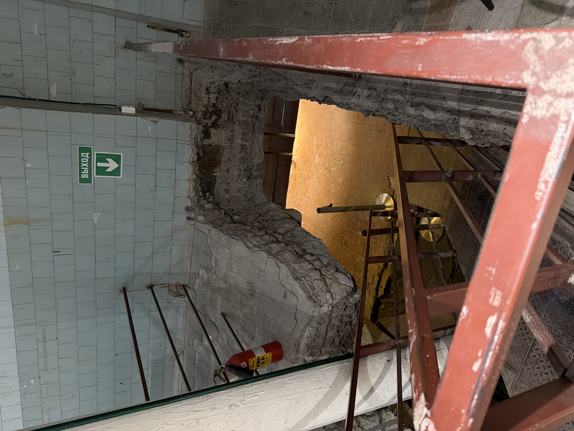 The image shows a construction site in Saint Petersburg, Russia. A hole has been created in a tiled wall, exposing a brick interior. A ladder provides access to the opening. A fire extinguisher is visible near the base of the wall. An "Exit" sign is present on the tiled wall. Metal scaffolding is in the foreground.