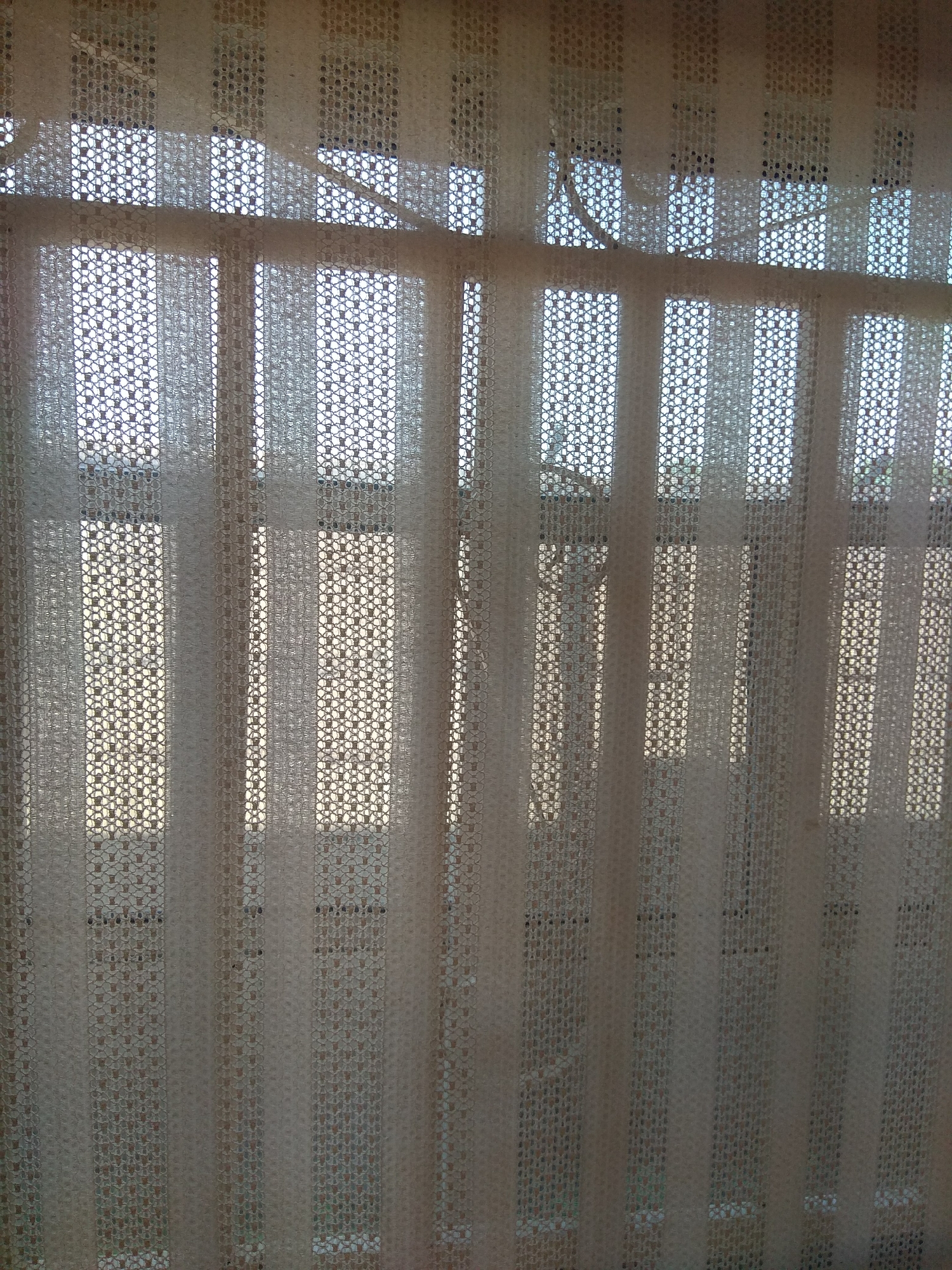 The image depicts a close-up view of a white, textured sheer curtain hanging in front of a window. The curtain has a repeating pattern of small, interconnected diamond shapes, creating a lace-like appearance. Light filters through the curtain and the window behind it, illuminating the textured surface and casting faint shadows. The window panes are visible through the sheer fabric, revealing a grid-like structure, possibly a window frame or blinds. The overall impression is one of diffused light and subtle detail, suggesting an indoor setting with natural light. The location in Jalingo, Nigeria, is inferred from the metadata associated with the image. There are no people or specific actions visible in the image, only the curtain and the light it filters.