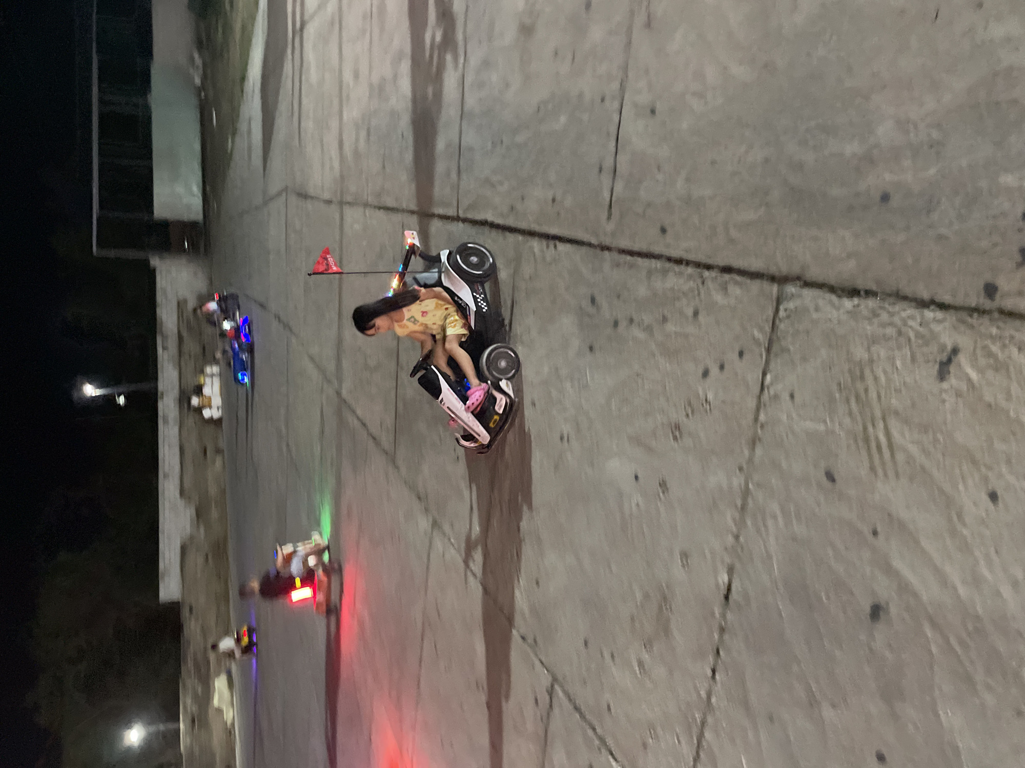 An image shows a young girl riding a go-kart at night in Mũi Né, Vietnam. The girl is wearing a yellow and orange dress and has long dark hair. She is sitting in a white and black go-kart with colorful lights on the back and a red flag on a pole. The go-kart has racing decals and a black steering wheel. The girl is holding the steering wheel and appears to be focused on driving. The go-kart is on a paved surface, possibly a race track or amusement park. In the background, other go-karts and people can be seen, with some of the vehicles illuminated by colorful lights. The scene is illuminated by artificial lights, likely from street lamps or the go-karts themselves, creating a dynamic and playful atmosphere. The overall impression is one of a fun evening activity at a tourist destination.