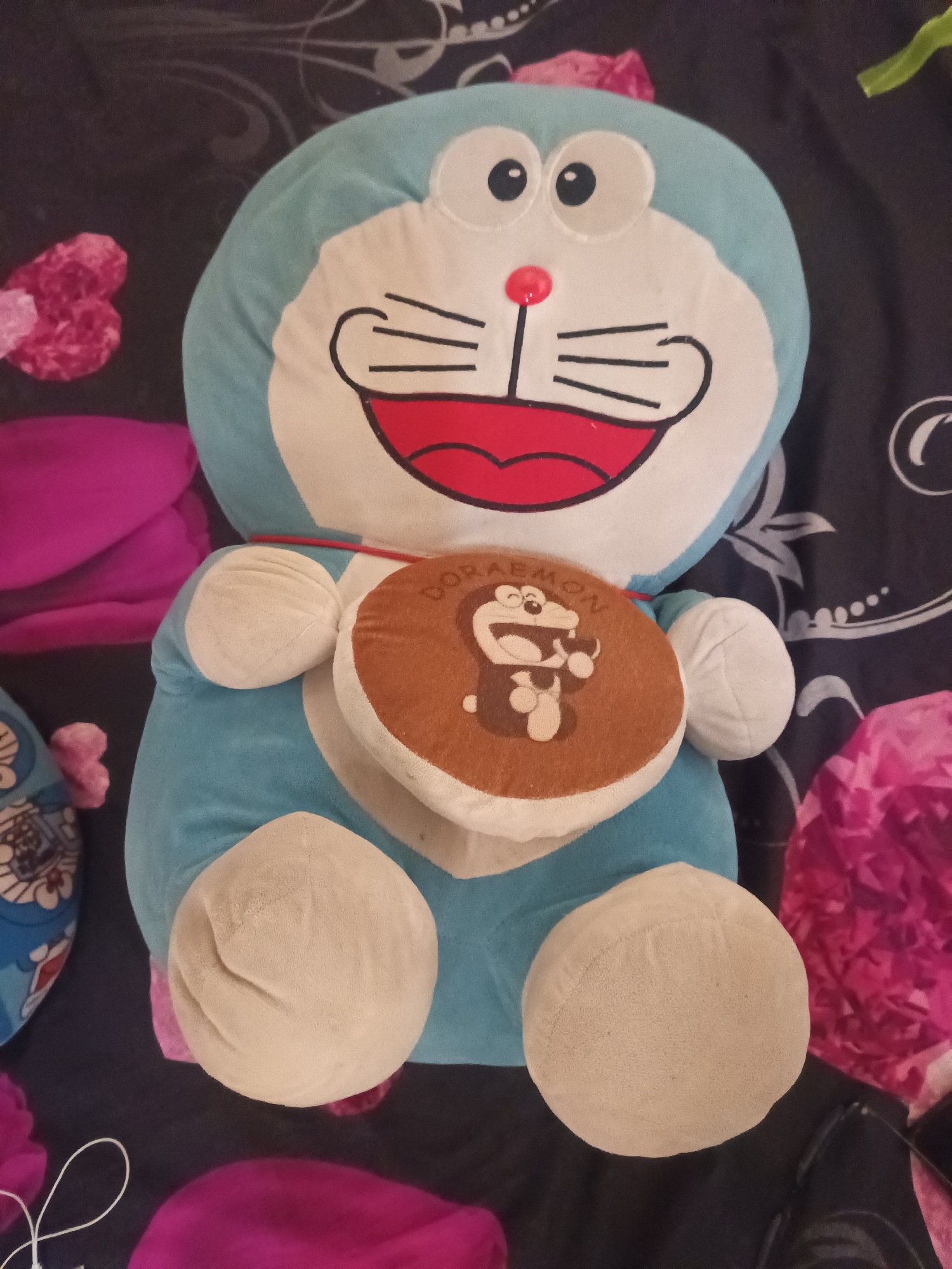 A plush toy of Doraemon, a blue robotic cat from the Japanese manga series, is situated on a dark, patterned surface, likely a bed or sofa. The toy is light blue with a white belly and face. It has large, round black eyes with white pupils, a red circular nose, and a wide, red mouth with black lines representing whiskers and teeth. A brown circular object, resembling a pancake or a bell with an image of Doraemon on it and the word "DORAEMON" embroidered in brown text, is attached to the toy's chest. The background features abstract floral patterns in pink and white. The lighting appears to be artificial and somewhat dim, creating soft shadows. The image is a close-up shot, focusing primarily on the Doraemon plush toy. There are no people visible in the image. The setting does not provide any specific clues about its location within Terbanggi Marga, Indonesia.
