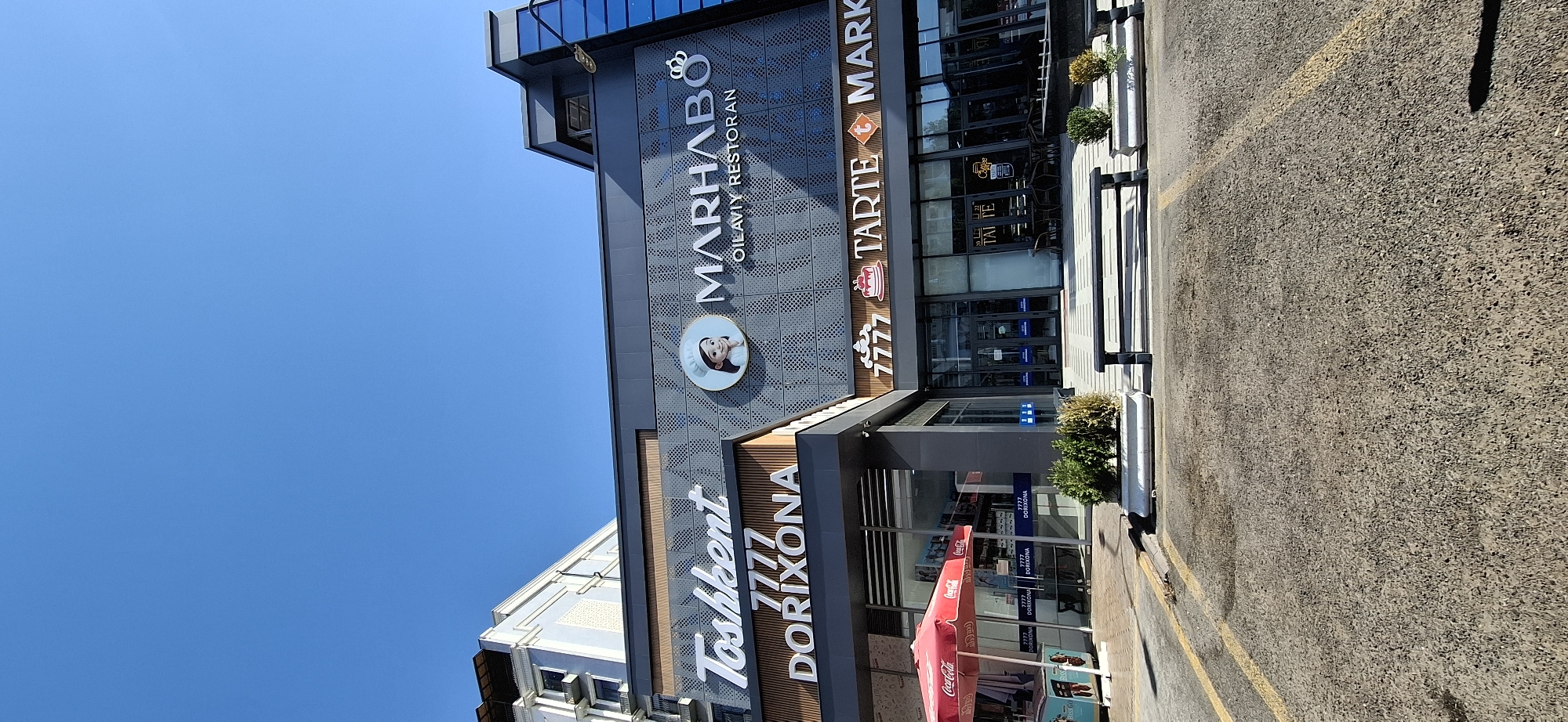 The image captures a sunny day in Tashkent, Uzbekistan, featuring the modern facade of a commercial building. The building displays prominent signage for "Toshkent 777 DORIXONA" (pharmacy) on the left and "MARHABO OILA VIY RESTORAN" (family restaurant) and "TARTE MARK" on the right. A red Coca-Cola umbrella is visible outside the pharmacy. The foreground shows a paved parking lot with yellow lines, and benches and planters with greenery are placed in front of the establishment. The sky is a clear, bright blue.