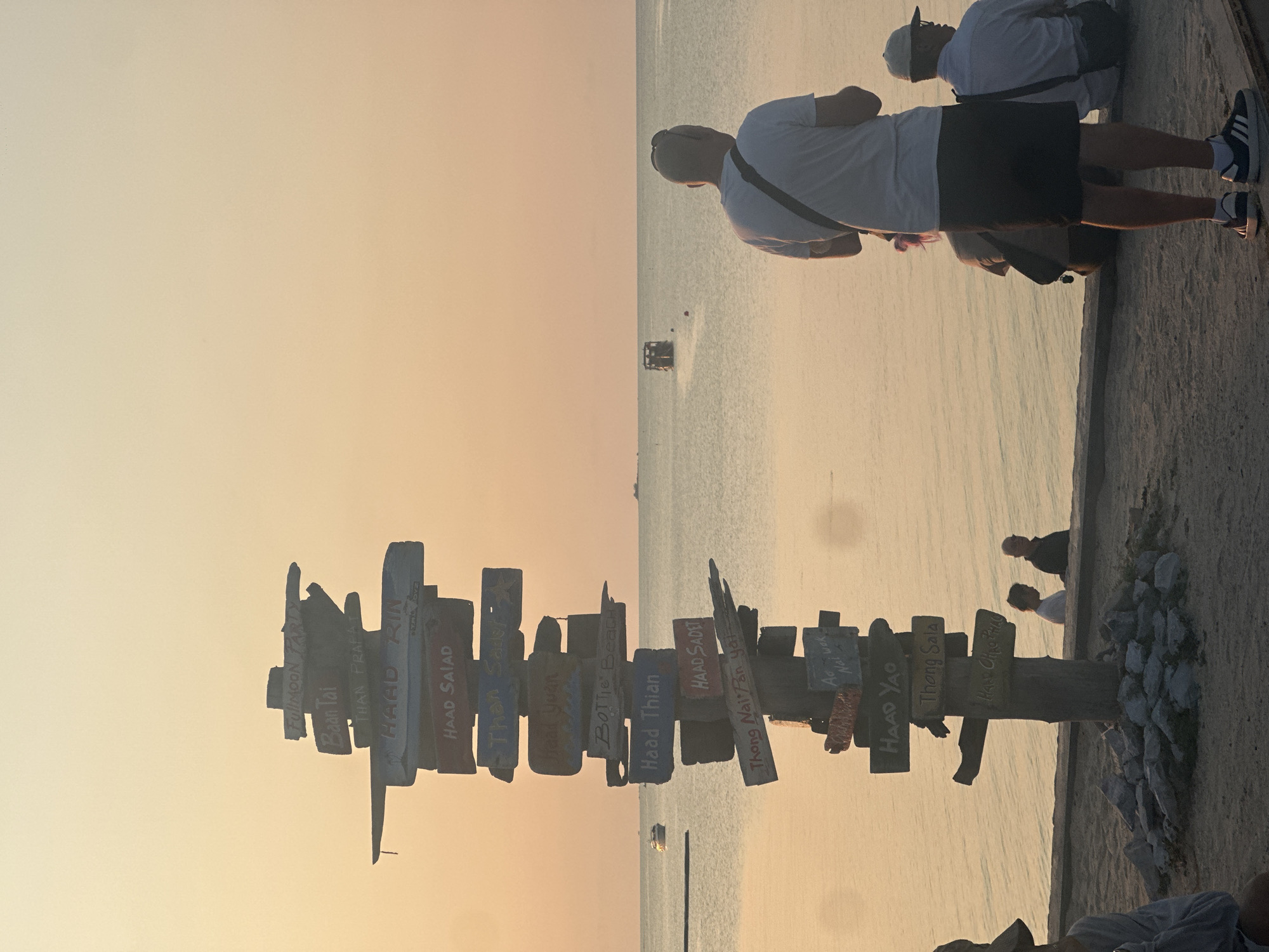 The image depicts a directional signpost on a beach, likely taken near sunset. The signpost is made of weathered wooden planks, each indicating different destinations. Destinations like "Fullmoon Party", "Baan Tai", "Haad Rin", "Haad Salad", "Haad Yuan", "Bottle Beach", "Haad Thian", "Haad Sadet", "Thong Nai Pan yai", "Haad Yao", "Thong Sala", and "Haad Ohao Phao" are visible on the sign. The post is surrounded by rocks.
In the background, there is a body of water, potentially the ocean, with a boat in the distance. The sky is a gradient of orange and yellow, indicating either sunrise or sunset.
On the right side of the image, there are people standing near the edge of what appears to be a concrete walkway. Some are looking out towards the water. The people are wearing casual clothing.
The image is taken in a way that it is flipped from its usual orientation. Given the OCR text, the location is likely near Baan Bang Rak, Thailand, based on the destination names written on the directional signpost.