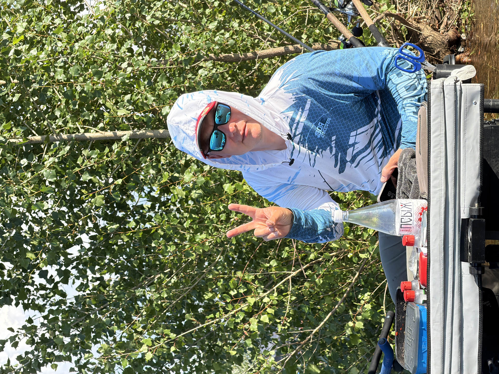 A man wearing a hooded fishing shirt and sunglasses is seated outdoors in Tarasovo, Russia, with his left hand raised in a peace sign. He is positioned behind a gray fishing tackle box, which contains various fishing accessories, including lures, a small box, and bottles. In front of the tackle box is a clear plastic bottle with a white label. Behind the man, lush green foliage from trees forms a backdrop. Two fishing rods are visible to the right of the man, leaning against a tree trunk. The man is facing forward, with a slight smile, and his sunglasses reflect the surrounding environment. The overall scene depicts a recreational fishing activity in a natural outdoor setting.