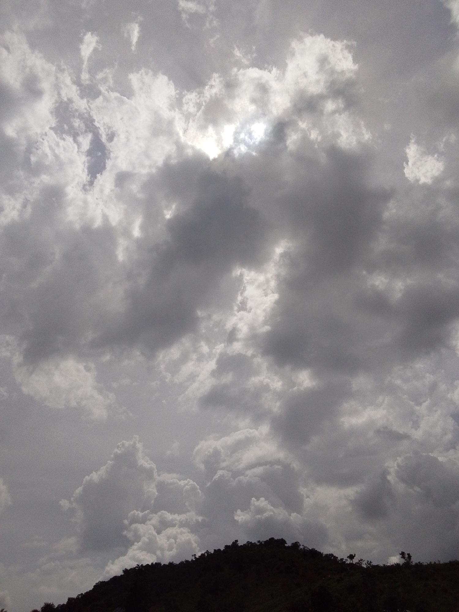 The image depicts a cloudy sky over a silhouetted hilltop, likely taken in Serti, Nigeria. The sky is filled with a variety of clouds, ranging from dark, heavy storm clouds to lighter, wispy formations. Sunlight is visible breaking through some of the clouds, creating bright patches. The hilltop is covered in dark foliage, with a few trees visible along the crest. There are no people or specific objects clearly discernible on the hilltop or in the sky. The overall impression is one of a dramatic and possibly impending weather event.