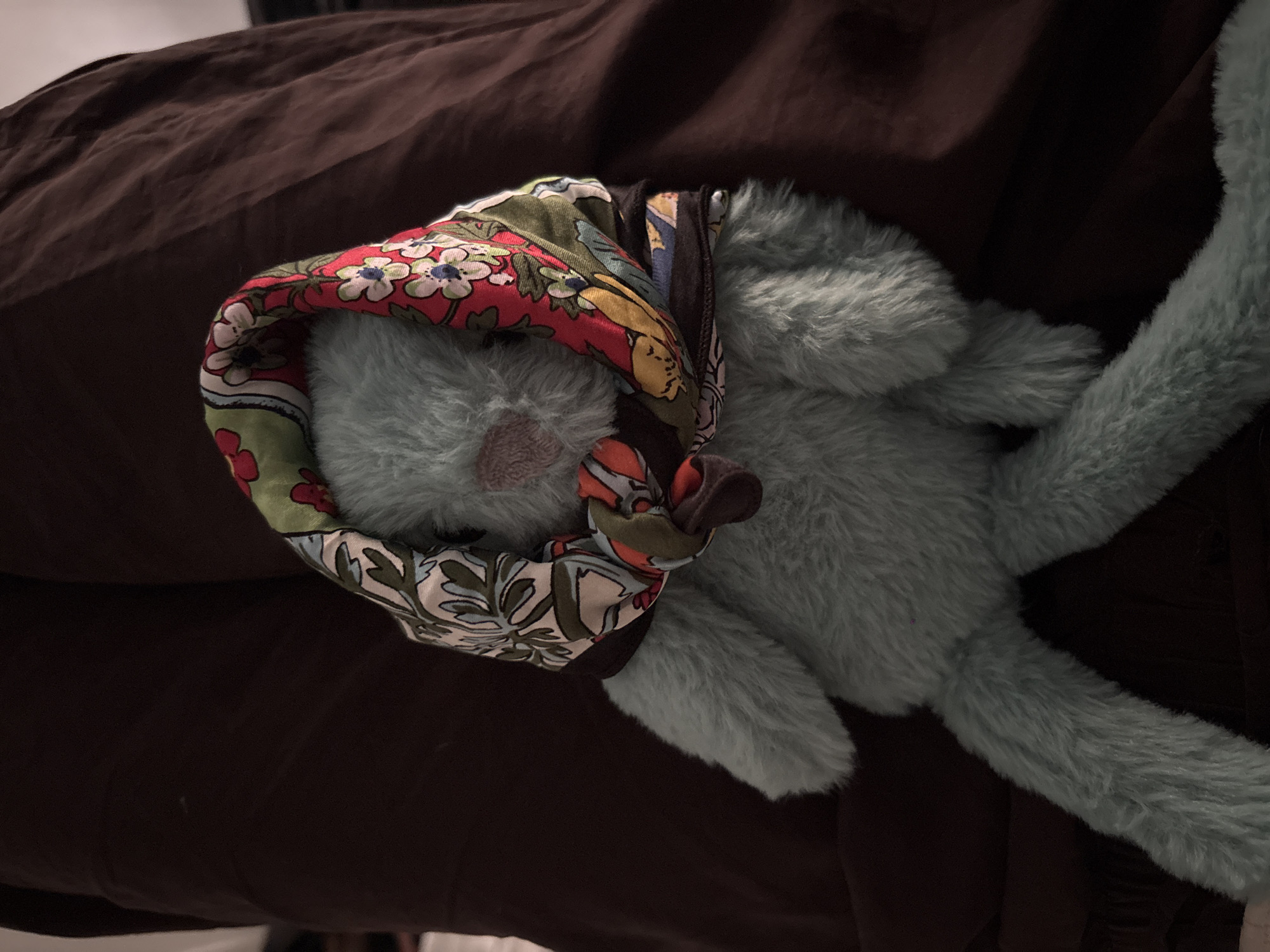The image features a light blue, fluffy stuffed animal, likely a rabbit or similar creature, lying on its back on a dark brown fabric surface. Its head is positioned towards the upper left, partially covered by a colorful, patterned scarf.
The scarf, tied around the stuffed animal's head in a babushka style, displays a vibrant floral pattern with prominent red, green, white, blue, and yellow hues. The stuffed animal's body extends diagonally across the frame, showcasing its soft, plush texture. The background consists of a dark brown, wrinkled fabric, possibly a blanket or bedsheet, providing a subdued contrast to the lighter-colored toy.
The scene depicts the stuffed animal in a state of rest. The lighting is dim and soft, suggesting an indoor setting, possibly in low light conditions or during the evening, contributing to a calm and peaceful atmosphere. There is no visible text in the image. While the location context is provided as Milan, Italy, the image itself contains no visual cues to indicate this geographical setting.