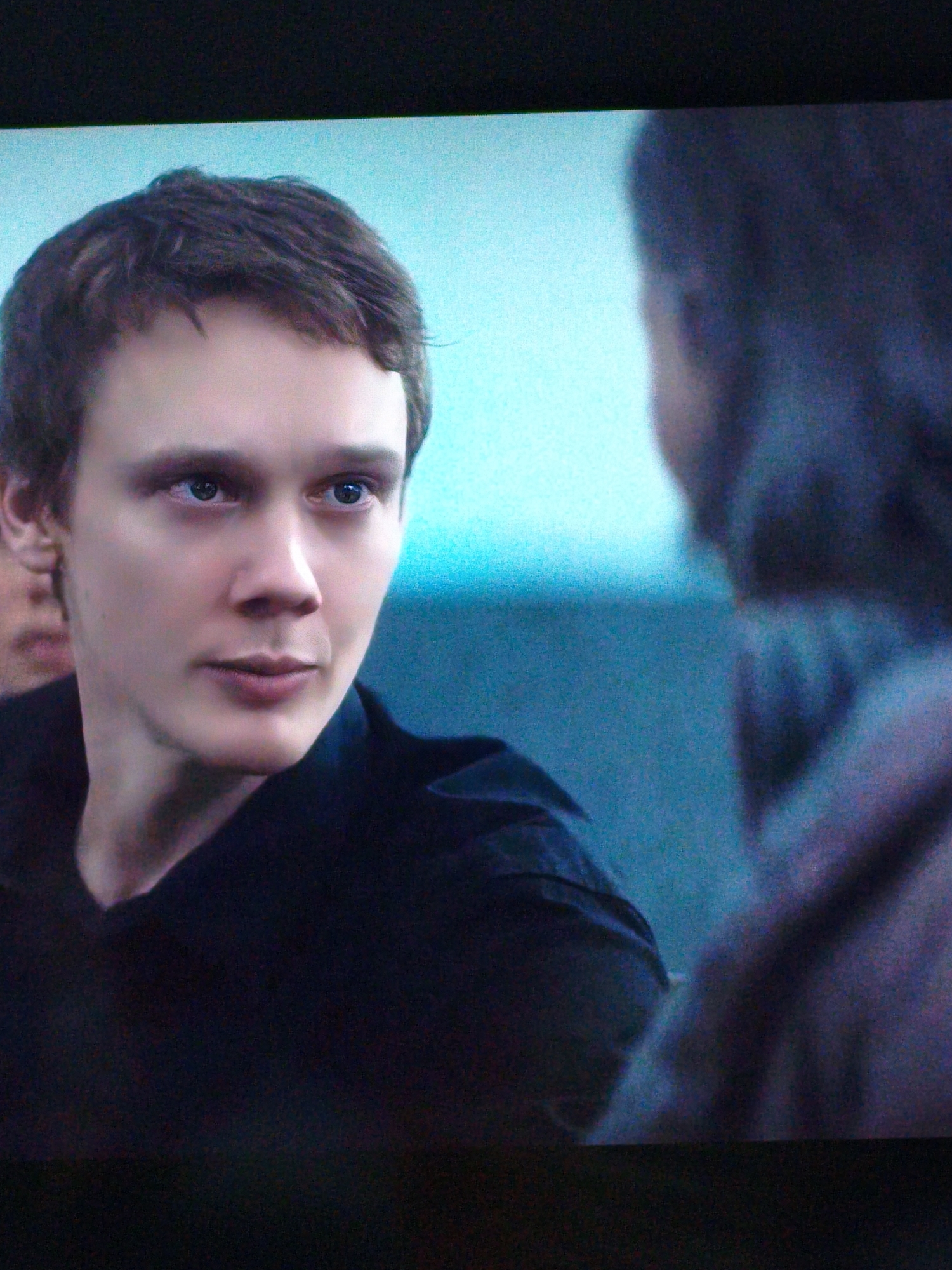 The image is a close-up, low-angle shot of a young Caucasian male with short, dark brown hair. He is wearing a dark, collared shirt. His gaze is directed to his left, towards another person who is mostly out of focus and visible in the right side of the frame. The background is a gradient of light blue to dark blue, suggesting an indoor setting with artificial lighting or a shallow depth of field. The overall lighting is dim and slightly grainy, typical of a screen capture or a low-light recording. There are no discernible elements that would indicate a specific location in Vladivostok, Russia.