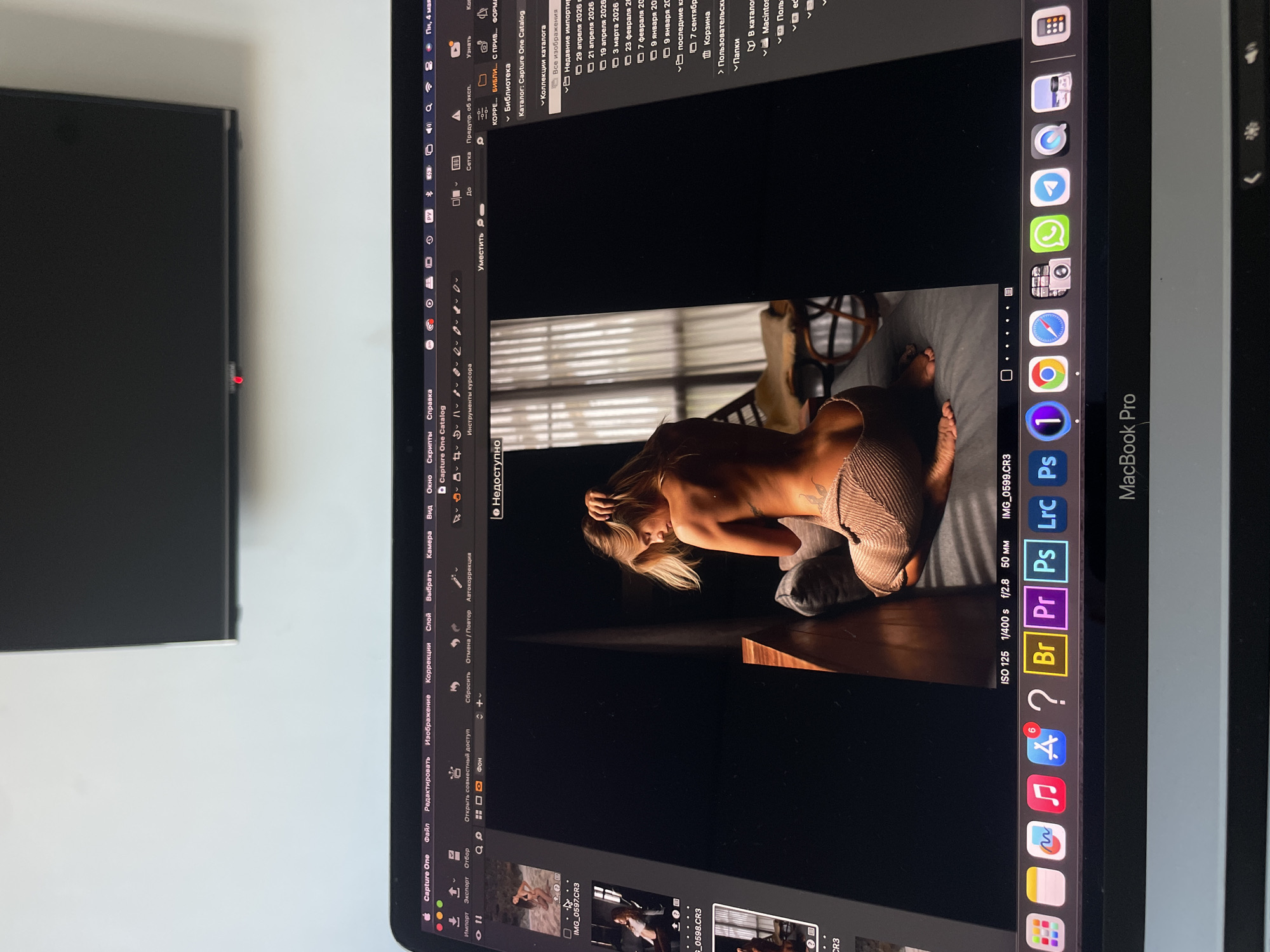 The image shows a MacBook Pro displaying the Capture One photo editing software. The software has an image of a partially clothed woman on the screen. The dock is visible on the right side of the screen with various application icons including Adobe Photoshop, Lightroom, and Bridge. The software interface language appears to be Russian. To the left of the Macbook is a TV screen. The background is a plain white wall.