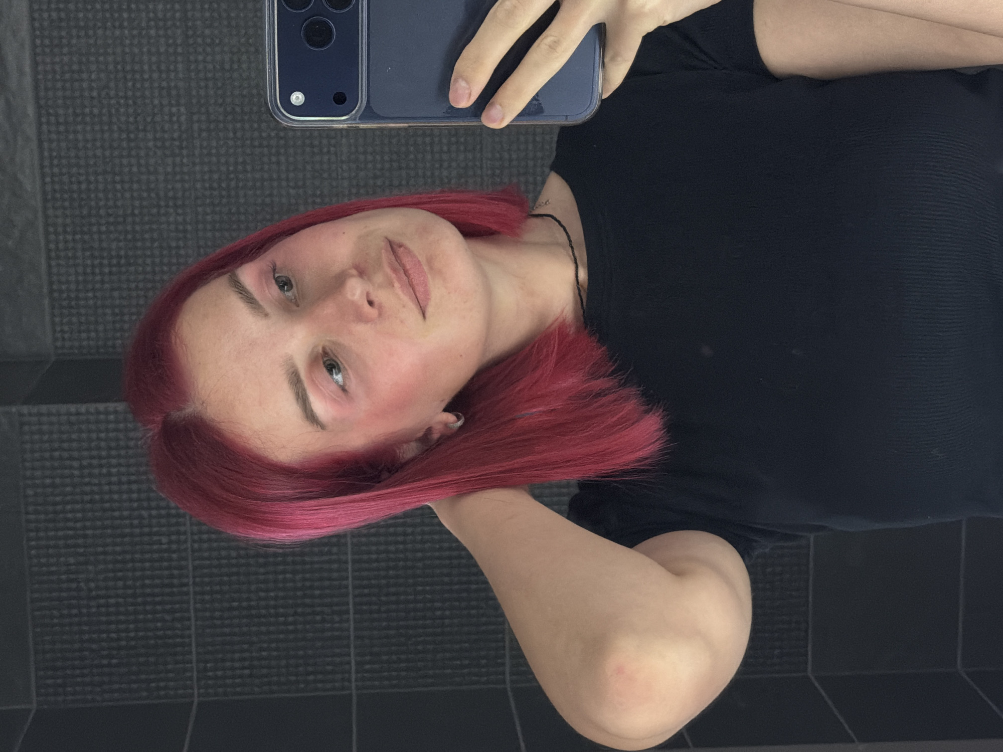 An indoor mirror selfie features a young woman with bright pink/red hair, captured in an image rotated 90 degrees counter-clockwise. The woman's head is positioned on the left side of the frame, tilted slightly, and she looks towards the camera with a neutral expression and a faint smile. Her eyes are light-colored, and she has a fair complexion with a subtle flush on her cheeks. She wears a black, ribbed short-sleeved top and a thin black necklace. Her right hand (viewer's left) holds a dark blue/grey smartphone with a clear case, its multi-camera module clearly visible. Her left arm (viewer's right) is bent, with her hand casually resting behind her head or hair. The background consists of dark grey tiles, some with a subtle grid-like texture, suggesting a bathroom or a similarly tiled room. No visible text is present in the image.