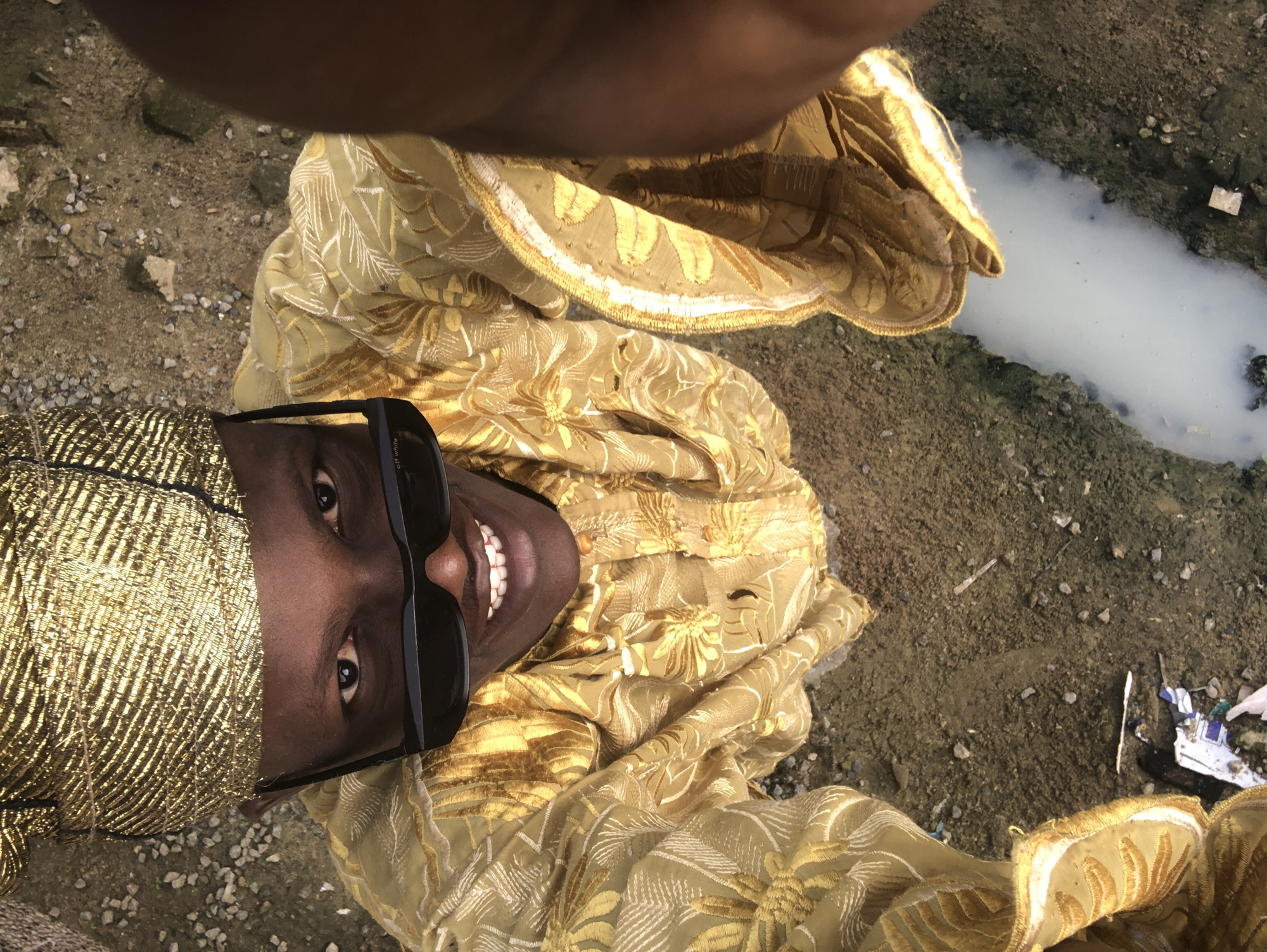 An upward-angle selfie taken from a high vantage point features a dark-skinned individual, appearing to be male, wearing a gold patterned kufi hat and a matching gold embroidered kaftan. The individual is also wearing black sunglasses and has a wide smile, revealing white teeth. The background is a rough, dry, dirt ground with scattered gravel and small debris. A narrow channel of murky, white liquid runs diagonally across the lower part of the frame, with some debris visible in and around it. The setting appears to be an outdoor, possibly urban or semi-urban, environment. The location is stated to be Ado, Nigeria.