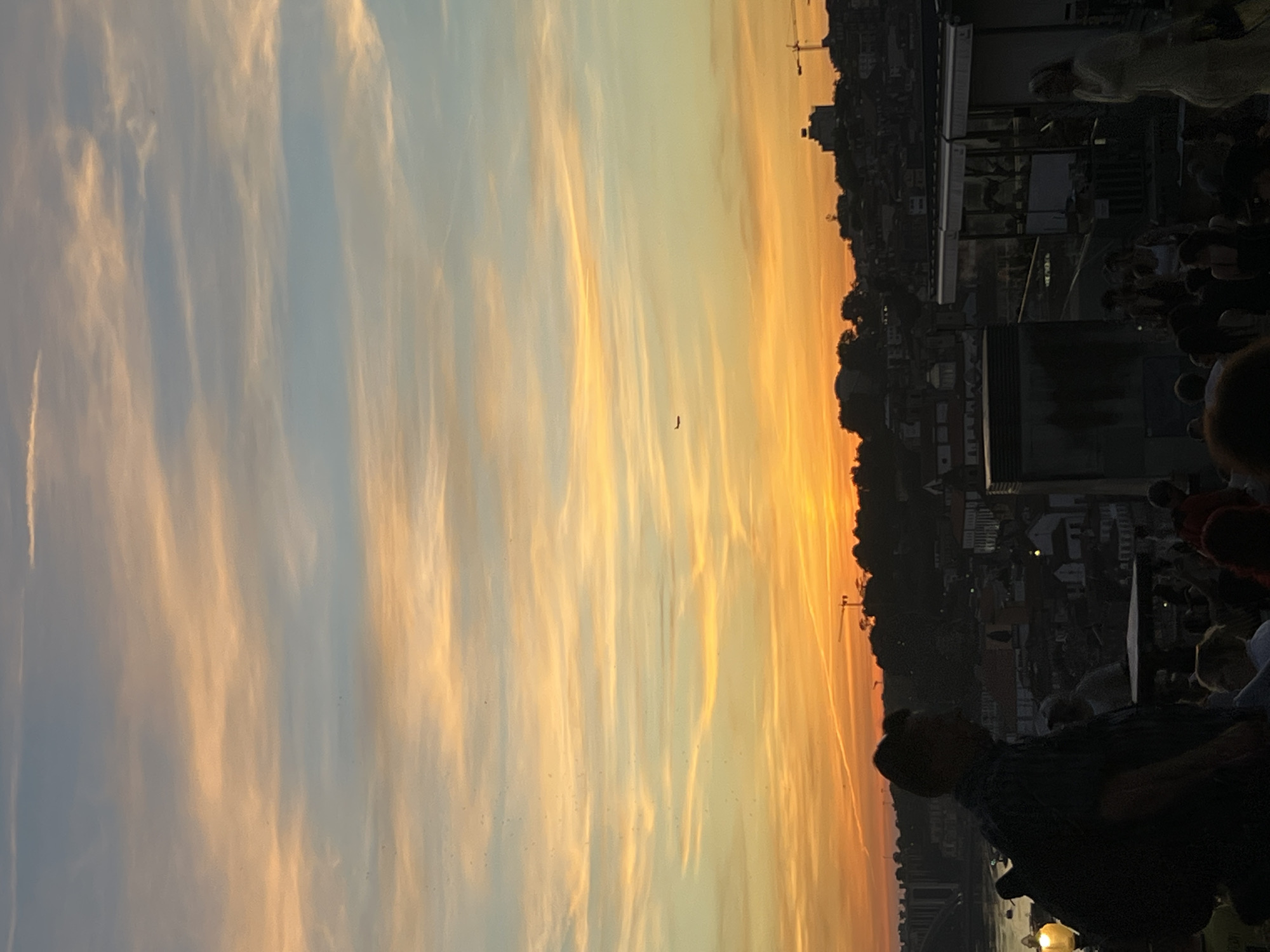 This image captures a sunset over the city of Vila Nova de Gaia, Portugal. The sky is a vibrant canvas of orange, yellow, and blue hues, with wispy clouds stretched across the horizon. A small airplane is visible in the distance. Below the sky, a silhouetted cityscape unfolds, with buildings and trees forming a dark outline against the colorful sky. Cranes are visible, suggesting ongoing construction. In the foreground, several people are gathered, their figures largely in silhouette due to the low light. One person, a male, is prominently featured on the left, facing away from the camera, wearing a backpack. Others are scattered throughout the scene, some appearing to be seated, others standing. The overall impression is of a bustling city enjoying the evening sky from an elevated vantage point.