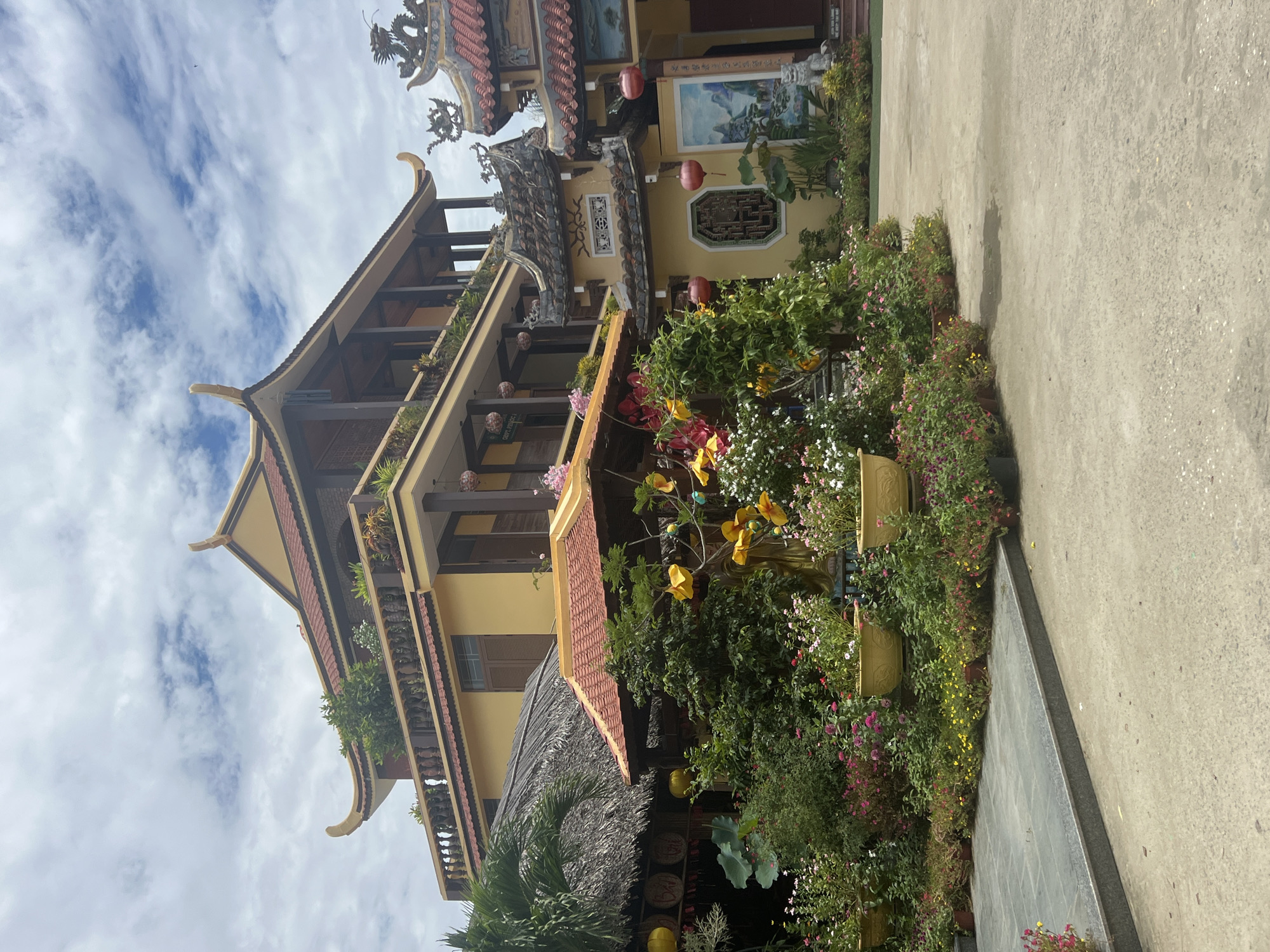 This is a photograph of a temple in Hoi An, Vietnam. The temple is a multi-story structure with yellow walls and red-tiled roofs in the traditional Vietnamese style. The building features ornate carvings and decorative elements, including dragon figures and lanterns. The foreground is filled with lush greenery, including a variety of flowering plants and trees in potted containers and beds. A paved courtyard leads to the temple entrance, and the sky above is cloudy.