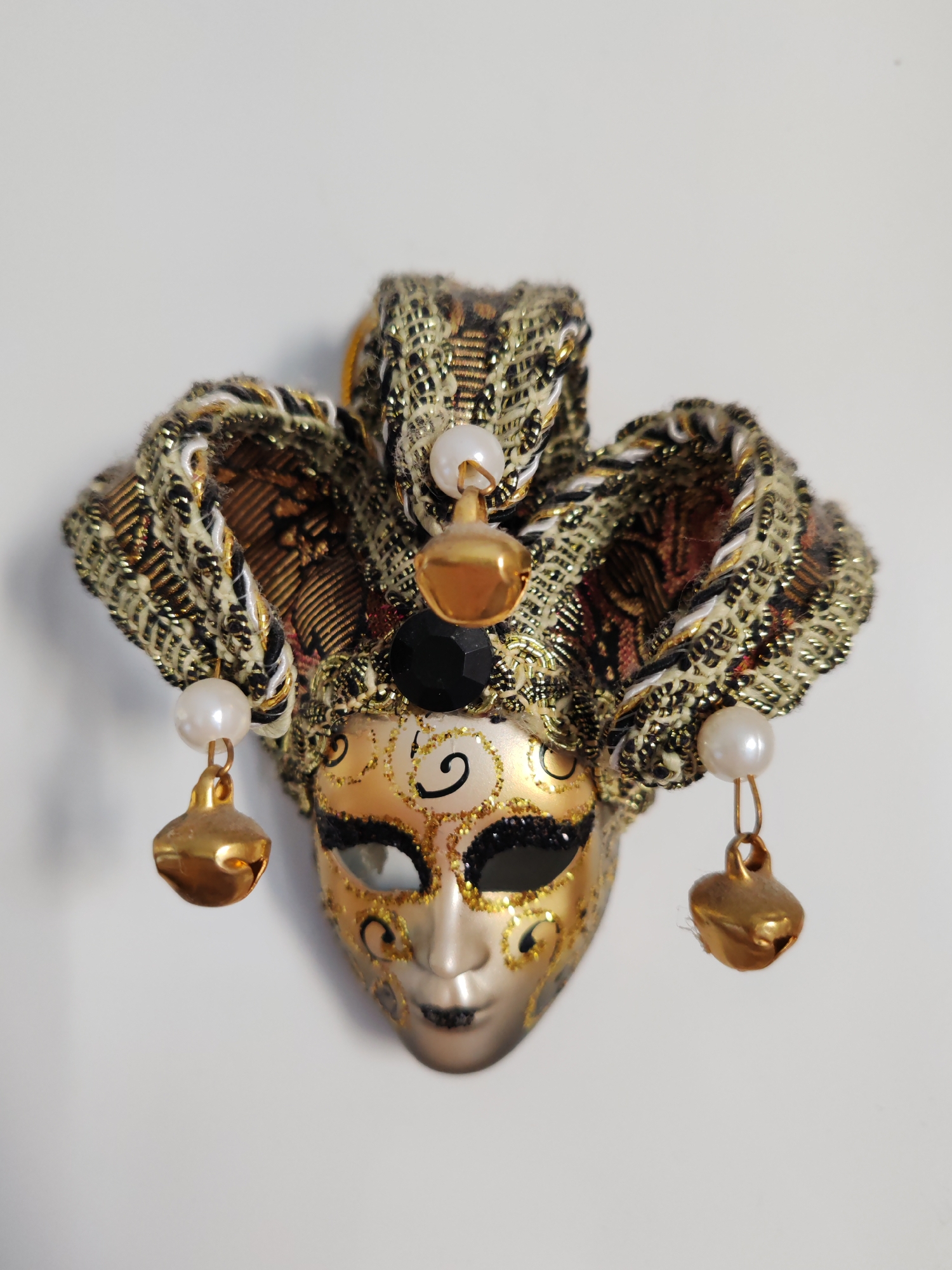 The image features a richly detailed Venetian-style jester or joker mask, displayed against a plain white background. The mask's face is predominantly gold and light beige, with one eye socket detailed in gold glitter and black paint, and the other in black glitter with gold accents. Intricate black and gold swirling patterns adorn the forehead and cheeks, while the lips are painted dark.
Above the mask, a three-pointed jester's hat is crafted from luxurious fabric, interwoven with gold, black, brown, and white threads, creating a shimmering, textured pattern. Each point of the hat is embellished with a creamy white pearl-like bead, from which dangles a small, polished golden jingle bell. A notable black faceted bead is positioned at the top center of the mask.
This is a still-life photograph, capturing the decorative object in detail. There is no visible activity or event, but the mask itself embodies the spirit of carnival, masquerade, and festive celebration. The lighting is soft and even, suggesting an indoor setting. No text is visible in the image. This ornate mask is observed in Saint Petersburg, Russia.