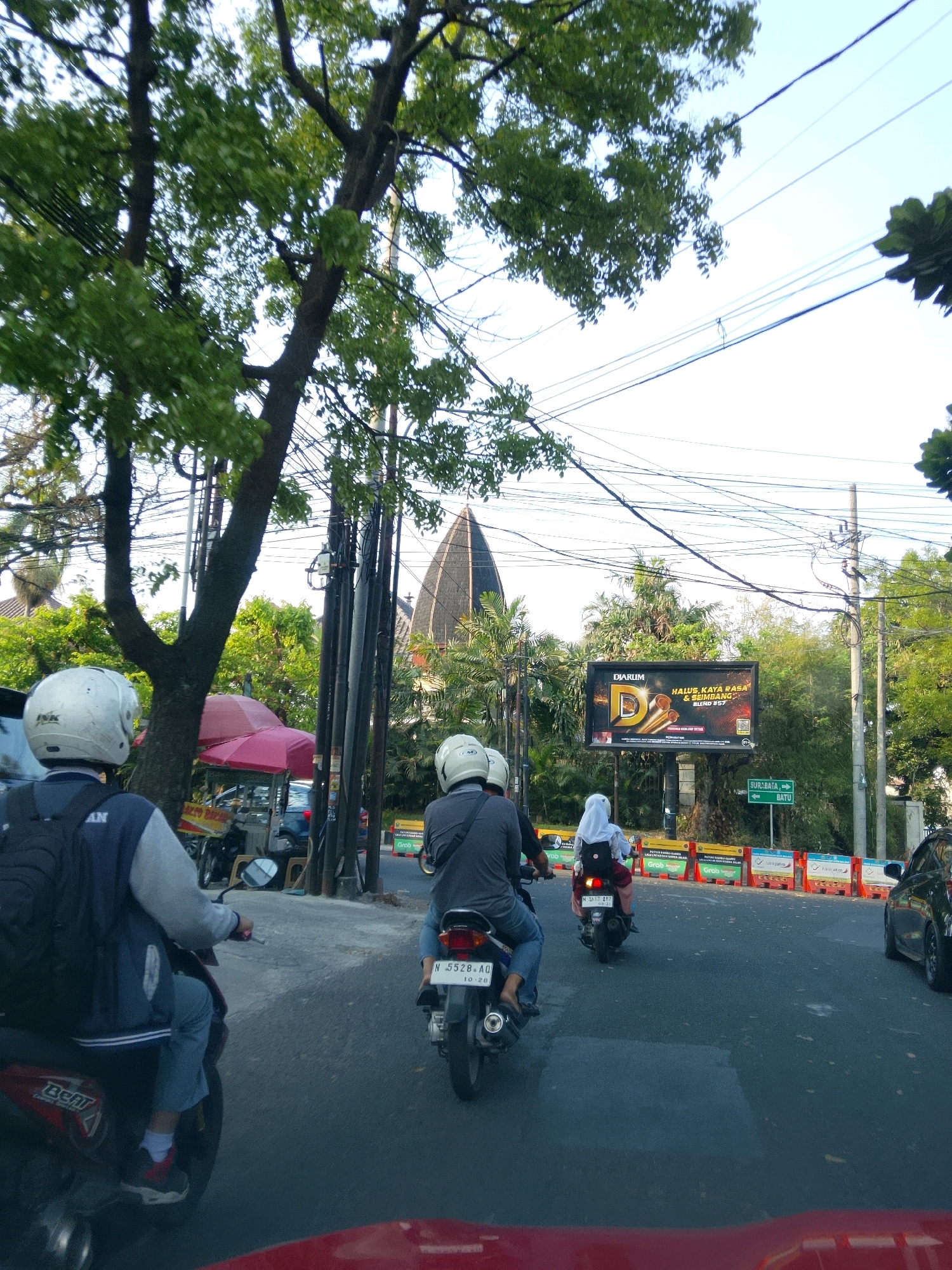 This is a photograph taken from inside a vehicle, looking forward at a street scene in Malang, Indonesia. The perspective is from a low angle, suggesting the photographer is seated in a car. The road is paved and appears to be a two-lane street.
In the foreground, on the left side of the image, is the back of a red vehicle, likely a car, with its roof visible.
To the left of the road, a tree with green leaves provides shade. Beside the tree, a food cart with a pink umbrella is visible, with some items on display.
Several motorcycles are on the road. In the immediate left foreground, a motorcycle is parked, with its rear end visible. Further ahead and slightly to the left, a motorcycle is being ridden by a person wearing a white helmet and a blue and grey jacket, carrying a black backpack. The motorcycle is red and black.
In the middle of the road, another motorcycle is being ridden by a person wearing a grey jacket and a white helmet. This motorcycle is black.
Further ahead and to the right, a third motorcycle is being ridden by a person wearing a white headscarf, suggesting it is a woman, and a red jacket. This motorcycle is black.
On the right side of the road, a black car is parked.
In the background, there are several poles with electrical wires strung between them.
A large billboard with a "DJARUM D" logo and Indonesian text is mounted on a pole. Below the billboard, a green directional sign indicates "SURABAYA" to the left and "BATU" to the right.
Beyond the road, there are many trees and some buildings. A distinctive building with a pointed, dark roof, possibly a mosque or a government building, is visible behind the trees. The sky is bright and clear.
The overall atmosphere suggests a sunny day in an urban or semi-urban environment.