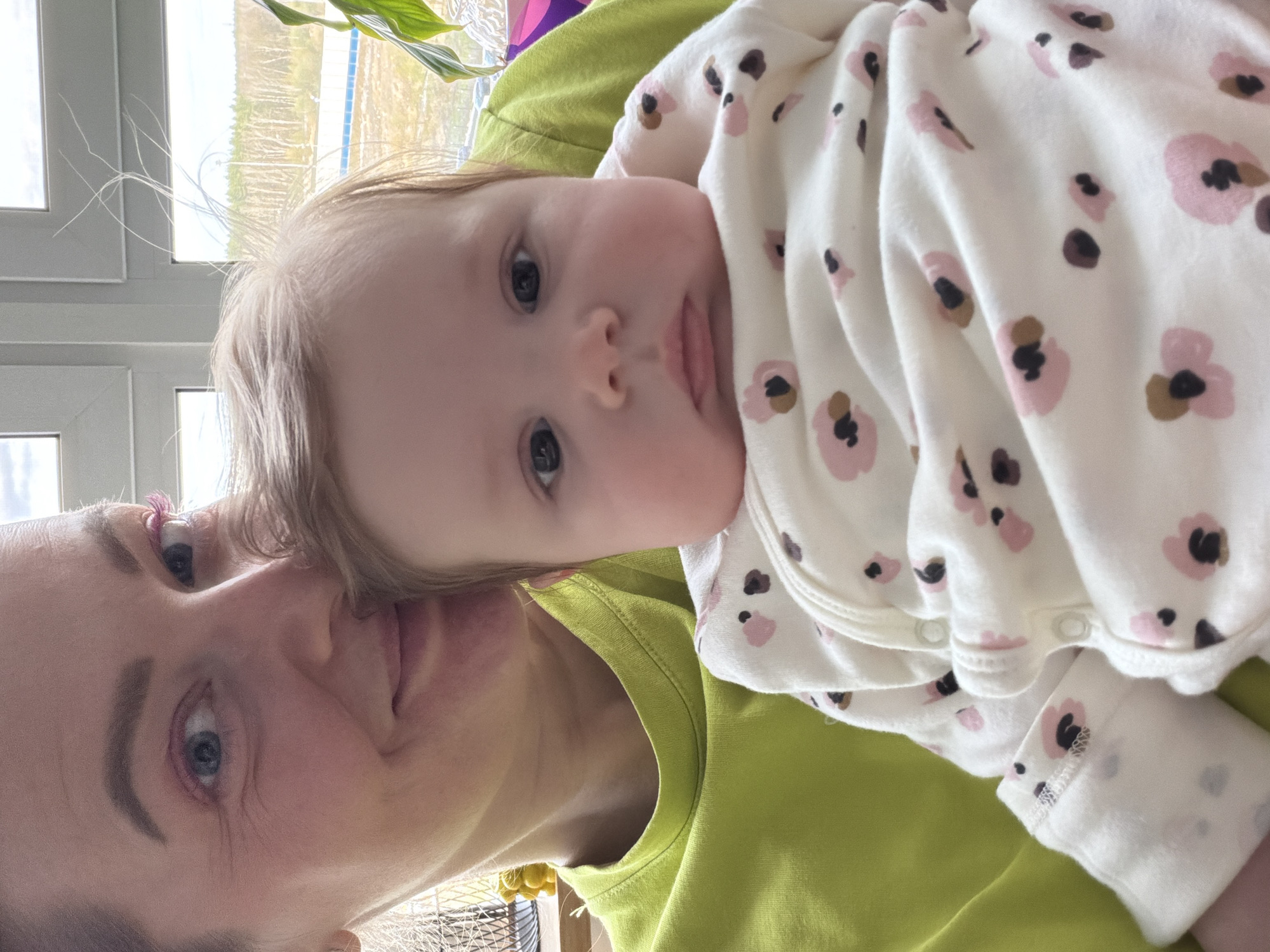 A fair-skinned adult with light blond hair and blue eyes holds a fair-skinned infant. The infant is lying on their back wearing a white onesie with pink and brown flower patterns. The adult is wearing a light green shirt. The setting appears to be indoors, with a window visible in the background. The geographic location is unknown.