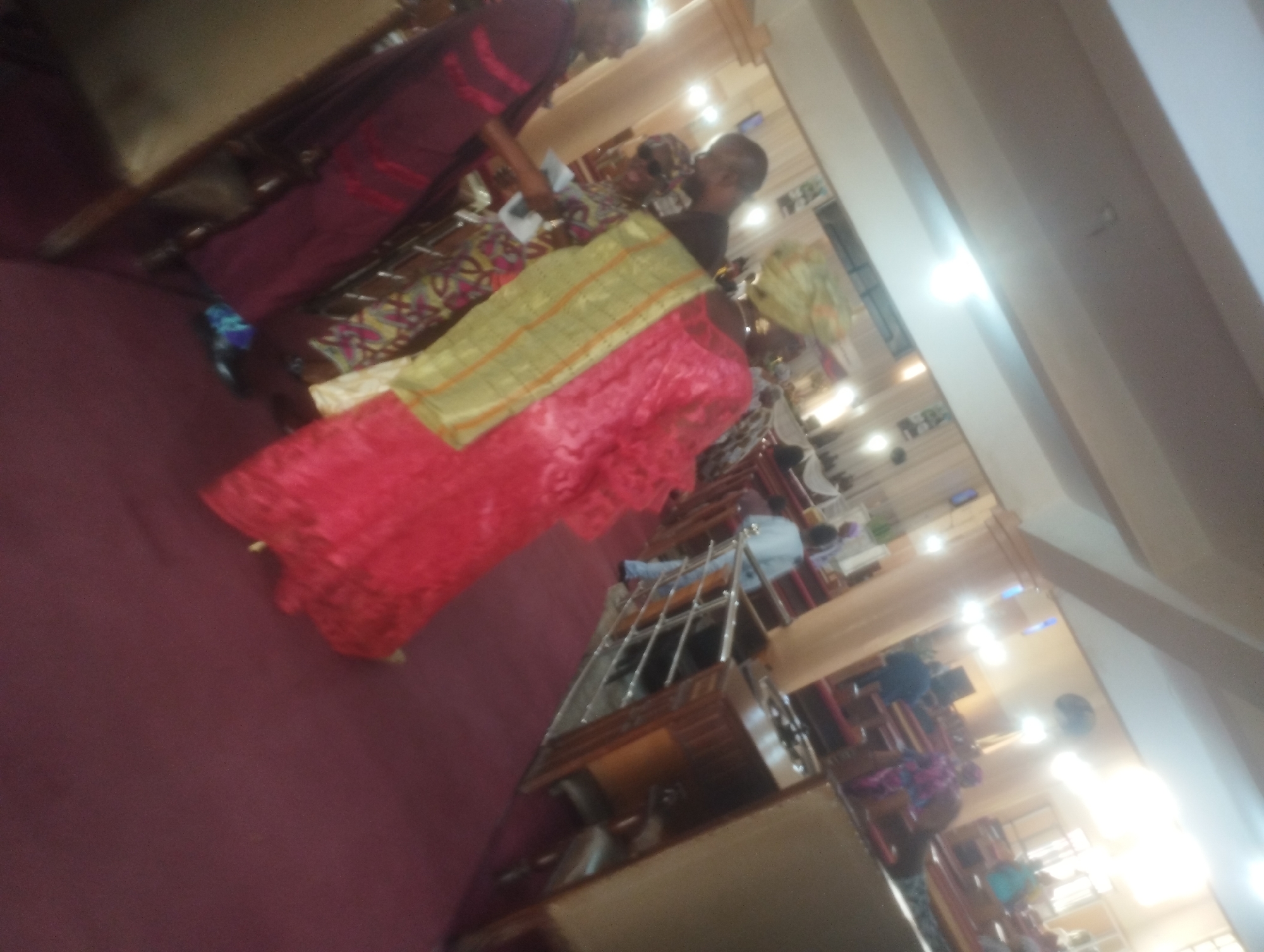 In an indoor setting that appears to be a place of worship or a gathering hall, likely in Jos, Nigeria, a group of people are present. The image is taken from a low angle and is quite blurry, making precise identification difficult. However, several individuals are visible, some dressed in traditional African attire. A prominent woman in the foreground is wearing a vibrant pink dress adorned with a gold and green sash or shawl. To her left, another person is seen in a maroon outfit. Further back, other individuals are partially visible, some seated or standing. The environment suggests an interior with wooden pews or seating, and lighting fixtures are visible on the ceiling, contributing to the overall illumination. The decor appears to be simple, consistent with a functional gathering space. The scene is tilted, indicating the camera was not held level.