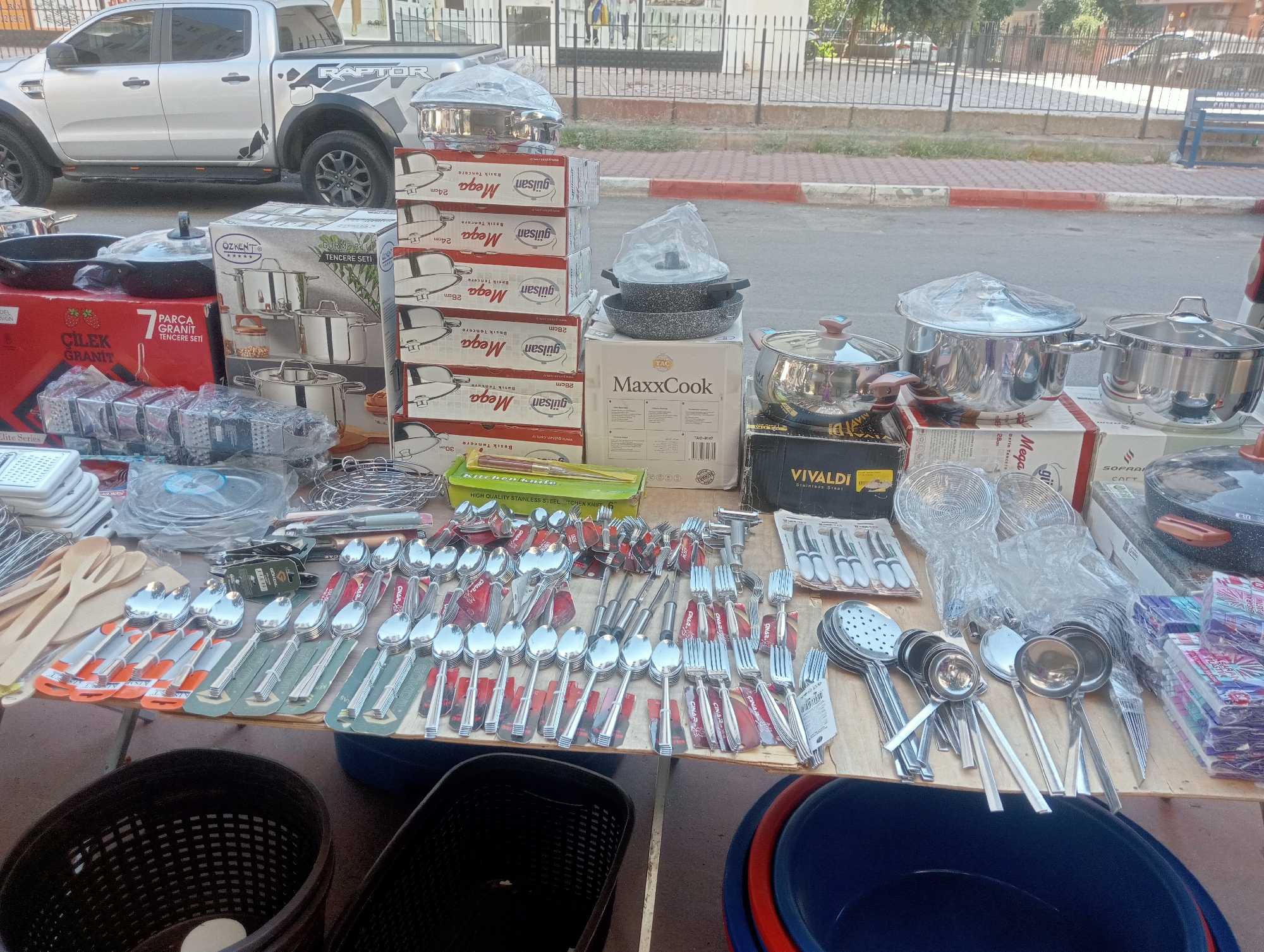 This is a street-level outdoor market scene, likely in Antalya, Turkey, featuring a variety of kitchenware for sale, predominantly cookware and cutlery. The backdrop includes a silver pickup truck with "RAPTOR" branding, parked on a street with a sidewalk, a metal fence, and buildings in the distance.
The foreground is dominated by a wooden table laden with merchandise. Prominently displayed are stacks of boxed cookware sets, including "Mega Tencere" (Mega Pot) and "MaxxCook" branded items. There are also individual pots and pans, some with plastic wrapping. A significant portion of the table is covered with an assortment of stainless steel cutlery, including spoons, forks, and serving utensils, neatly arranged in packaging. Wooden kitchen utensils and kitchen knives are also visible.
Several plastic storage baskets, one dark brown and partially filled, and another black, are positioned at the bottom of the frame. To the right, large blue and red plastic basins are visible.
The overall impression is one of a bustling street vendor setup, showcasing a wide array of kitchen essentials. The lighting suggests it is daytime. No people are clearly visible in the immediate foreground, though figures can be seen in the distant background.