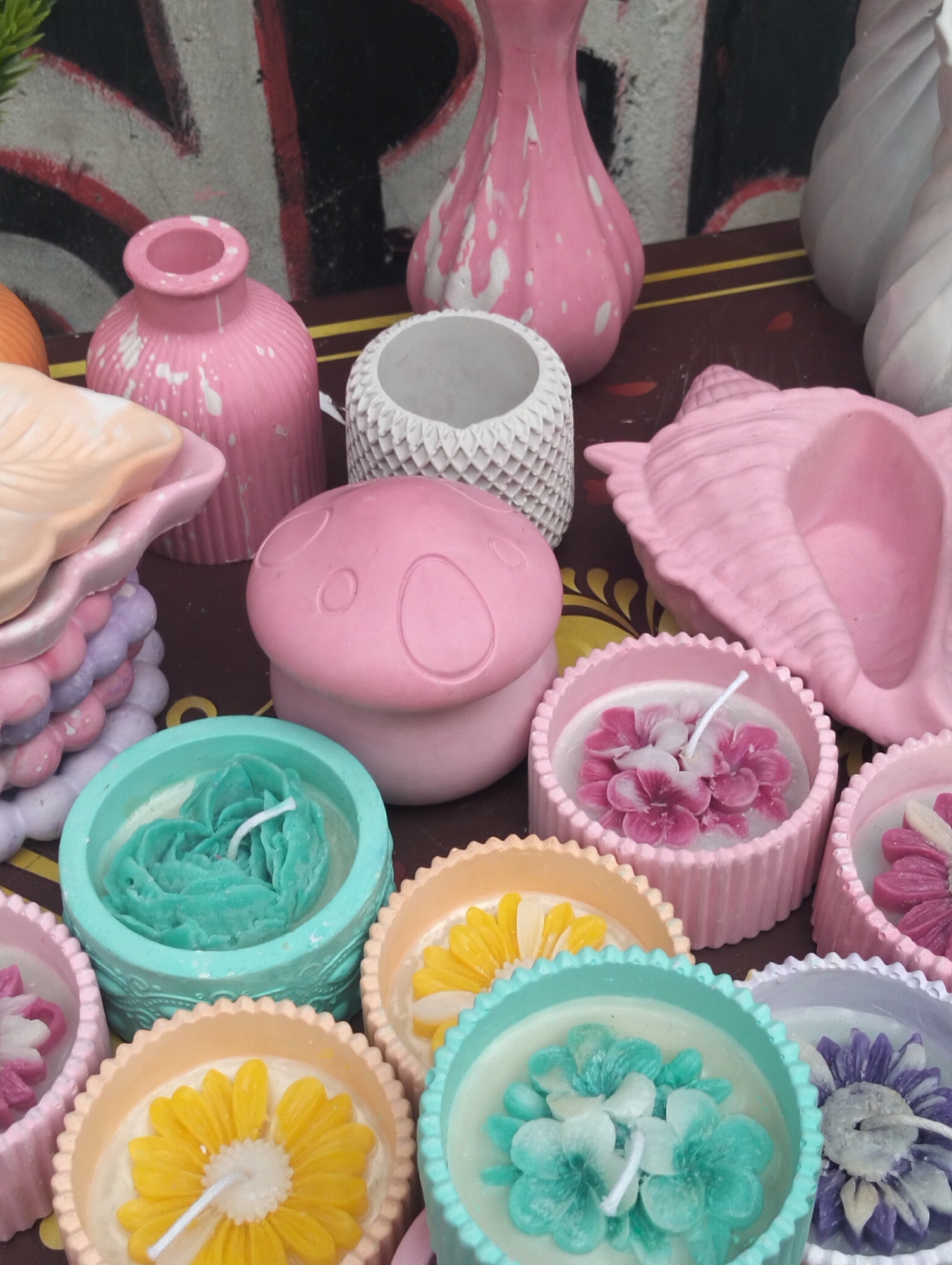 The image depicts a tabletop display of decorative items, including vases, small containers, and flower-shaped candles in pastel colors. The candles are arranged in circular containers with scalloped edges, and the flowers are white with colorful tips. The vases and containers are made of a similar material, possibly ceramic or plaster. The background shows a wall with graffiti or painted designs. The location is identified as Dhaka, Bangladesh.