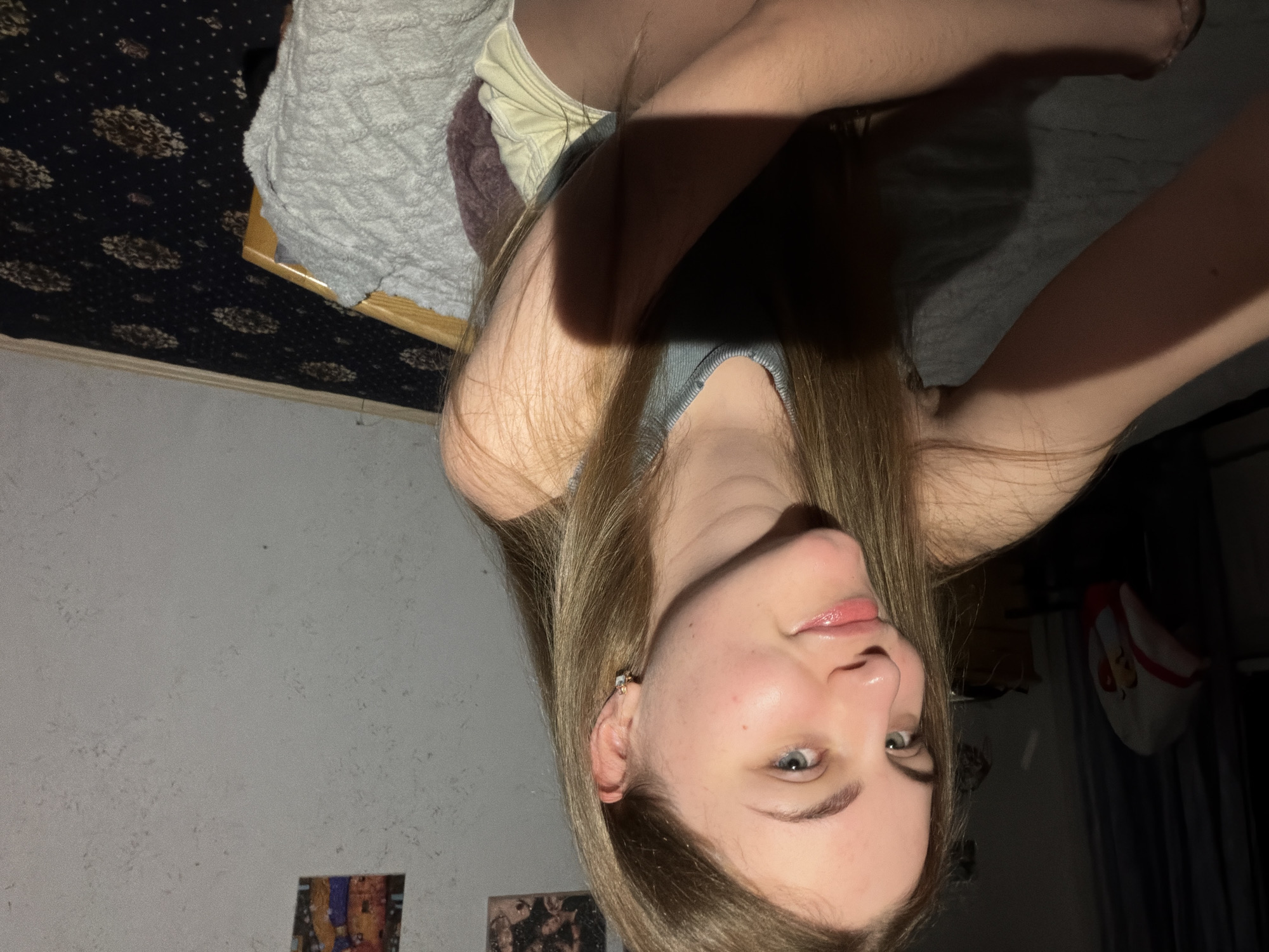 A young person with long blonde hair, wearing a grey tank top, lies on a bed. They are looking up and to the right, with a slight smile and their eyes open. Their right arm is raised, with their hand resting near their head. A shadow from their arm falls across their chest and face. The room is dimly lit, suggesting it might be evening or night. In the background, a dark carpet with a floral pattern is visible on the left, and a white wall with some framed pictures can be seen on the lower left. To the right, a dark curtain and a bag with a cartoon character are visible. The overall mood is relaxed and casual.