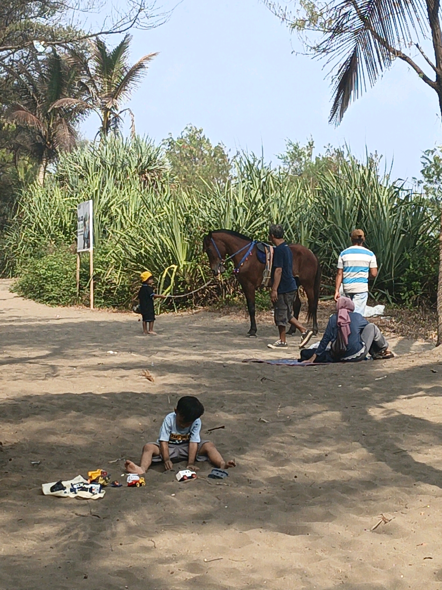 In Tambakmulyo, Indonesia, a sandy area is depicted with several people and a horse. In the foreground, a young boy sits on the sand, playing with several small toy vehicles. To the right of the boy, a person wearing a hijab and a dark blue top is seated on the sand, facing away from the camera. Behind this seated person, another individual wearing a blue and white striped shirt and a brown cap stands, facing away. Between these individuals and the horse, a man in a dark blue t-shirt and shorts walks. A brown horse, saddled, stands to the left of the man. Further to the left, a child wearing a yellow helmet and a dark outfit stands, holding a stick. A signpost is visible behind this child. The background consists of tall, green vegetation and palm trees, under a clear blue sky. The ground is predominantly sand with scattered debris.