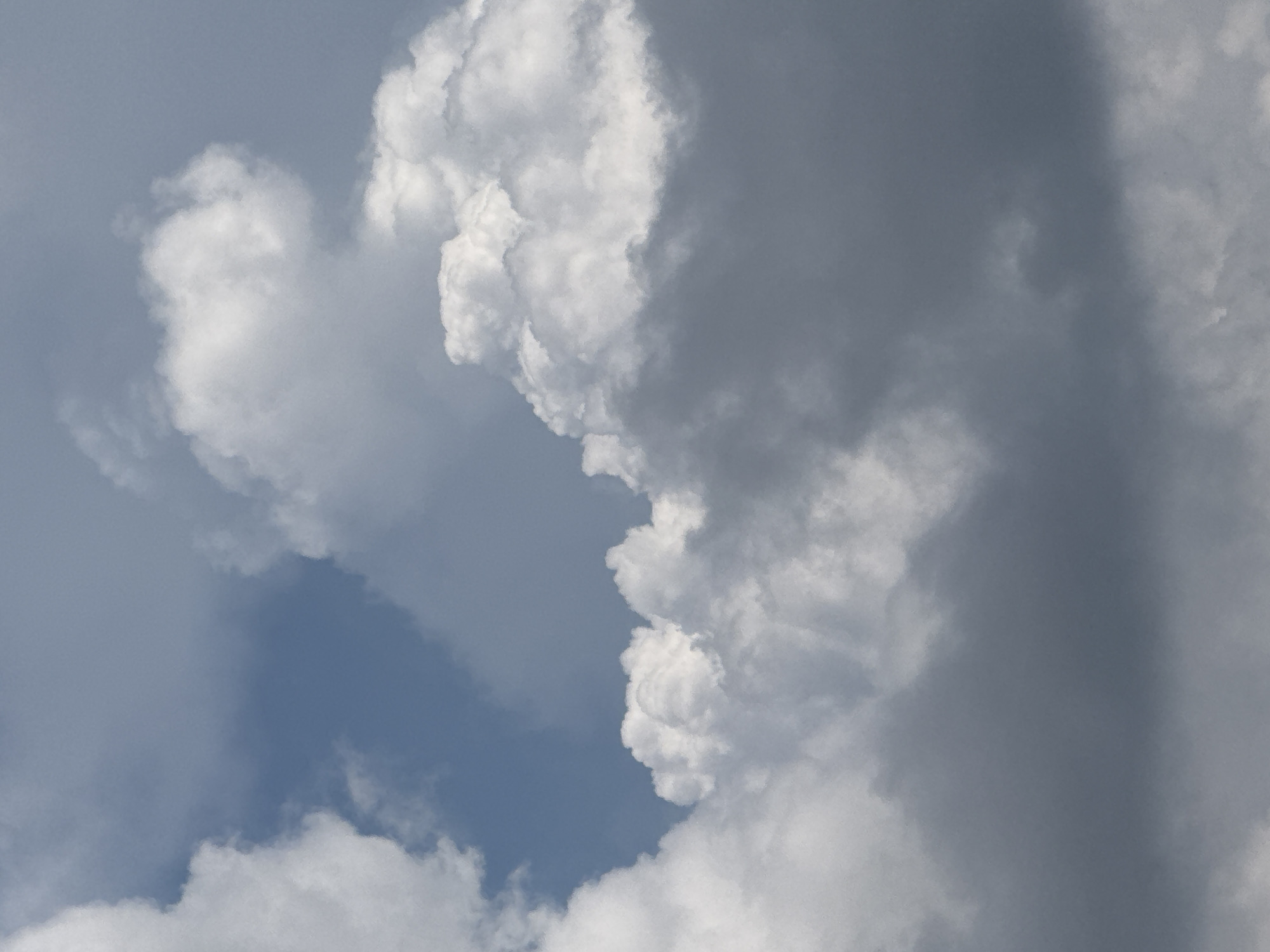 The image shows a cloudy sky. Cumulus clouds, some bright white and others dark gray, fill the frame. A patch of blue sky is visible towards the upper left. The image does not depict any specific location in the city of Đa Lộc, Vietnam, nor does it contain any people, subjects, objects, or actions. It is a natural atmospheric scene.