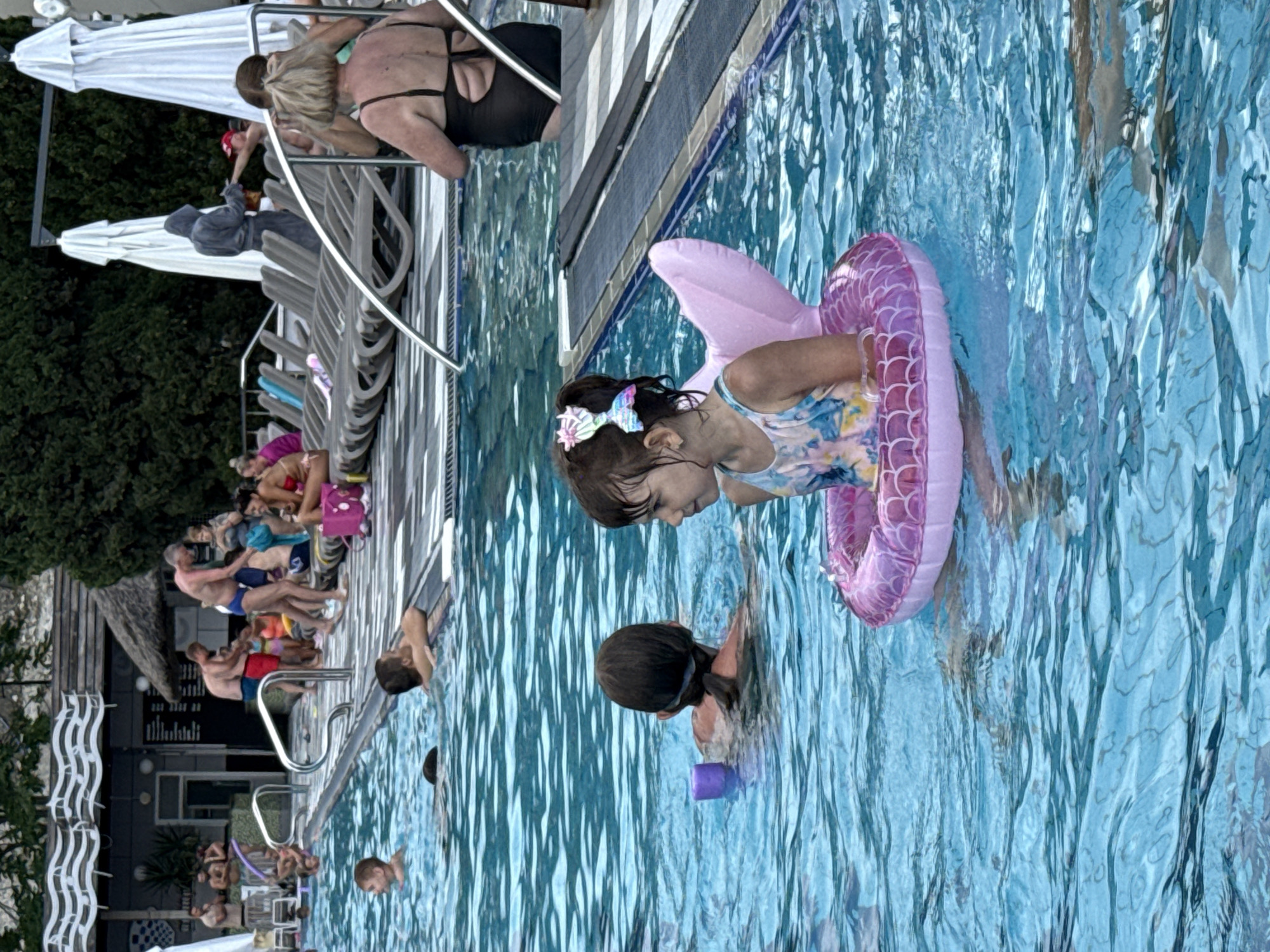 The image depicts a sunny day at an outdoor swimming pool. In the foreground, a young girl with a butterfly hair clip and a mermaid-themed floatation device is immersed in the blue water. To her left, another child, viewed from behind, is also in the pool with a purple float. The background shows a scene with several adults and children around the pool, some lounging on chairs, others standing or swimming. The environment appears to be a recreational area with trees and umbrellas visible.
