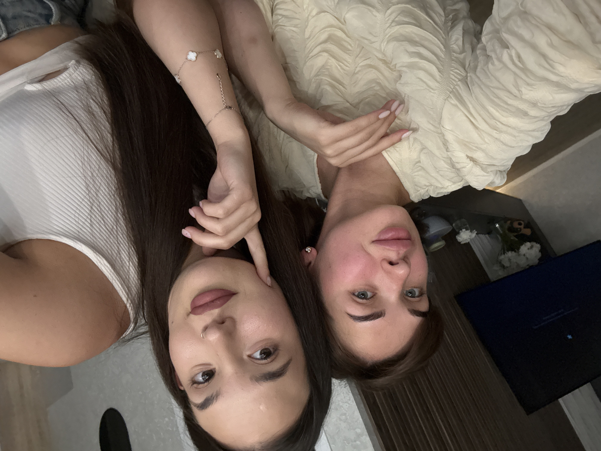 Two women are closely positioned, looking at the camera, which appears to be held at an unusual angle. The woman on the left has dark hair, a nose ring, and is wearing a white ribbed top. The woman on the right has lighter hair and is wearing a textured cream-colored top. Her fingernails are painted a light pink color. A bracelet is visible on her left wrist. In the background, there is a dark TV screen, a flower bouquet, and a light-colored floor, indicative of an indoor setting. It is not possible to confirm the location as Лобаново, Russia.