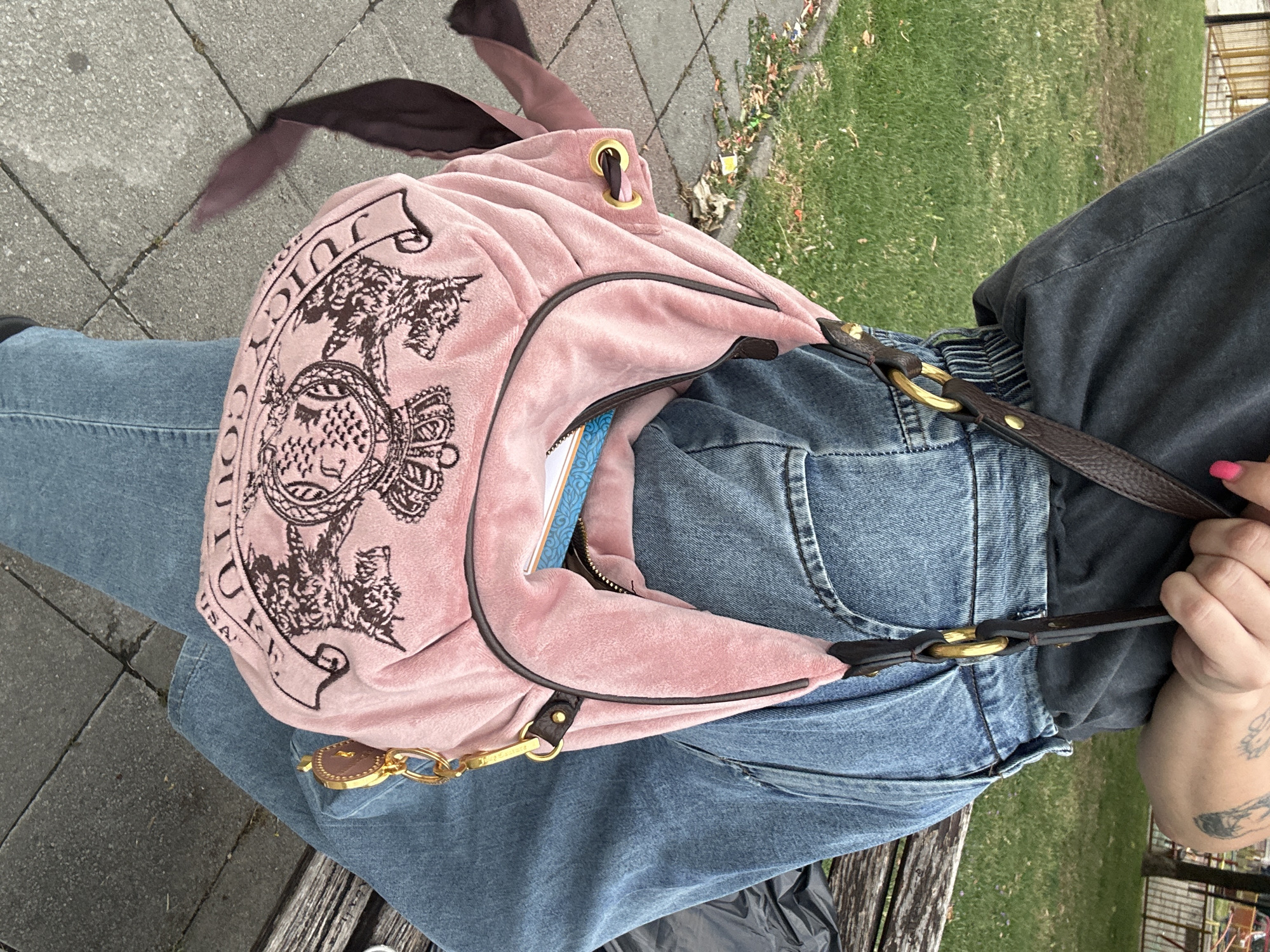 A person wearing a dark gray t-shirt and loose-fitting blue jeans is sitting on a park bench. They are holding a light pink velour shoulder bag with brown trim and gold hardware. The bag has a crest logo with two dogs and a crown. A small brown charm hangs from the strap. The background shows a paved walkway with some leaves, a grassy area, and a blurred fence. The scene is outdoors in what appears to be a public park in Belgrade, Serbia.