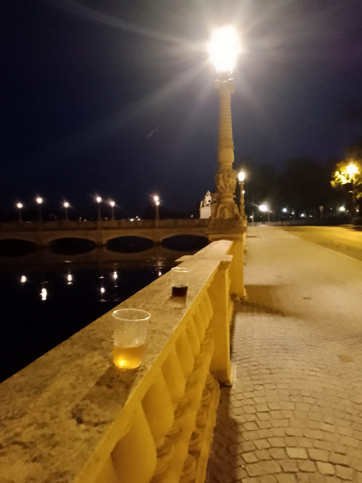 A nighttime, eye-level, medium shot shows two clear plastic cups, one with a light brown liquid, and the other with a dark brown liquid, resting on a stone railing. The railing is part of a bridge with ornate yellow balusters. Beyond the railing, a dark body of water reflects the lights of a bridge in the distance, which has multiple lampposts. A tall, ornate lamppost stands to the right of the frame, its light casting a strong glare. A cobblestone walkway stretches into the background, illuminated by streetlights. The sky is dark blue and clear. This scene is located on the Pfaffenteich bridge in Schwerin, Germany.