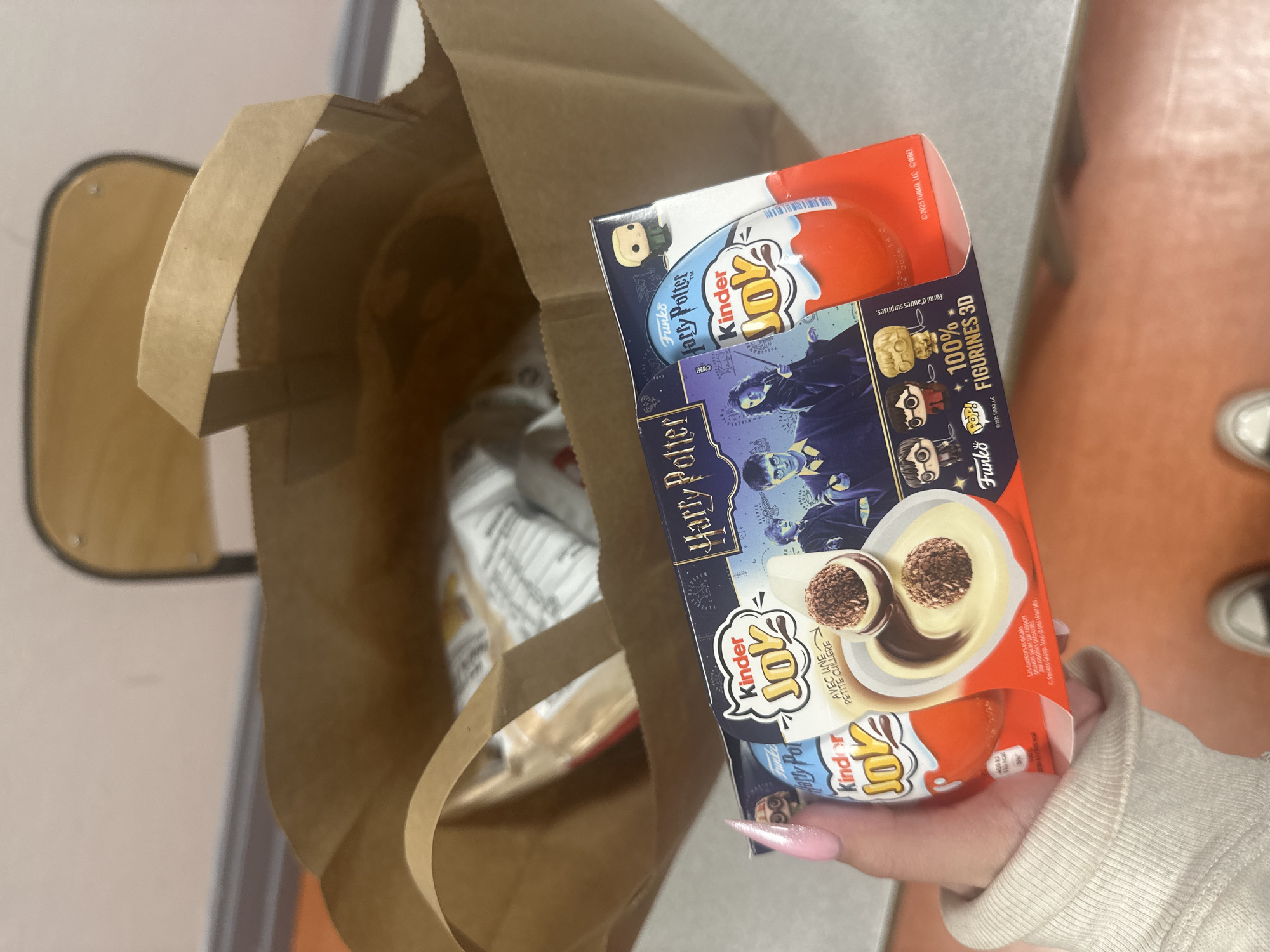 The photograph shows a person holding a box of Kinder Joy Harry Potter Edition chocolate candy. The box features images of Harry Potter characters and the Kinder Joy logo. The person's hand is visible, with pink nails. Next to the box is a partially visible brown paper shopping bag, which appears to contain other items. The background is out of focus but suggests an indoor setting, possibly a kitchen or eating area with a counter and flooring. The image was taken in Strasbourg, France.