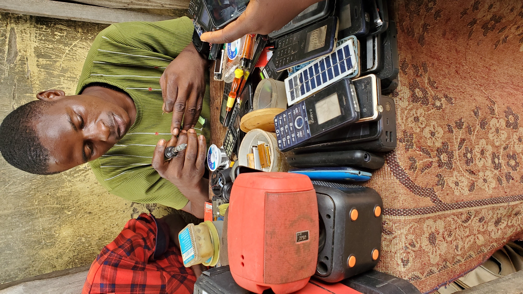 A young Nigerian man in a green ribbed shirt is working on electronic devices, likely repairing them. He is seated at a table covered with a patterned rug, surrounded by various mobile phones, phone parts, tools including a soldering iron, wire, and screwdrivers. A red speaker and a black power bank are also visible on the table. In the background, a portion of another person in a red and black checkered shirt is seen. The setting appears to be an outdoor stall or workshop, with a textured, weathered wall behind the man. The scene is indicative of a mobile phone repair business operating in Jalingo, Nigeria.