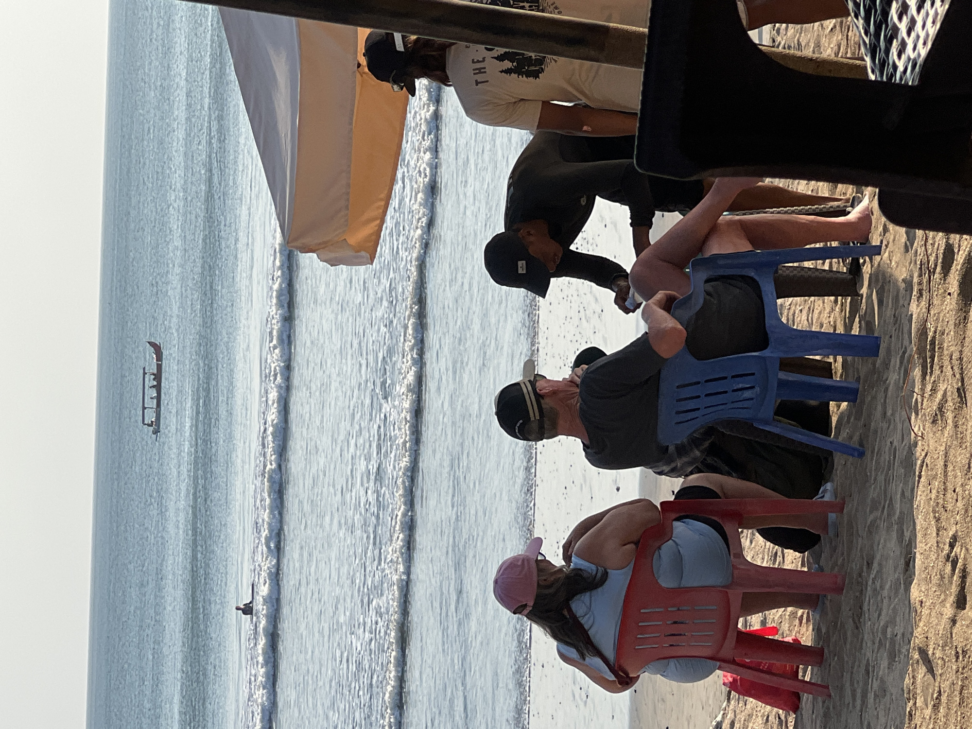 This image depicts a beach scene in Kuta, Indonesia, under a bright sky. In the foreground, two individuals are seated on chairs facing the ocean. To their right, two other people are engaged in an interaction. One, wearing a black wetsuit and cap, appears to be handing something to the other person who is wearing a light-colored t-shirt with text and an image on the back. The beach sand is visible beneath them. In the midground, the ocean stretches out with gentle waves rolling onto the shore. A surfer is visible in the water, and further out, a boat with several passengers is navigating the sea. The scene is framed by parts of a beach umbrella or canopy in the upper right corner.