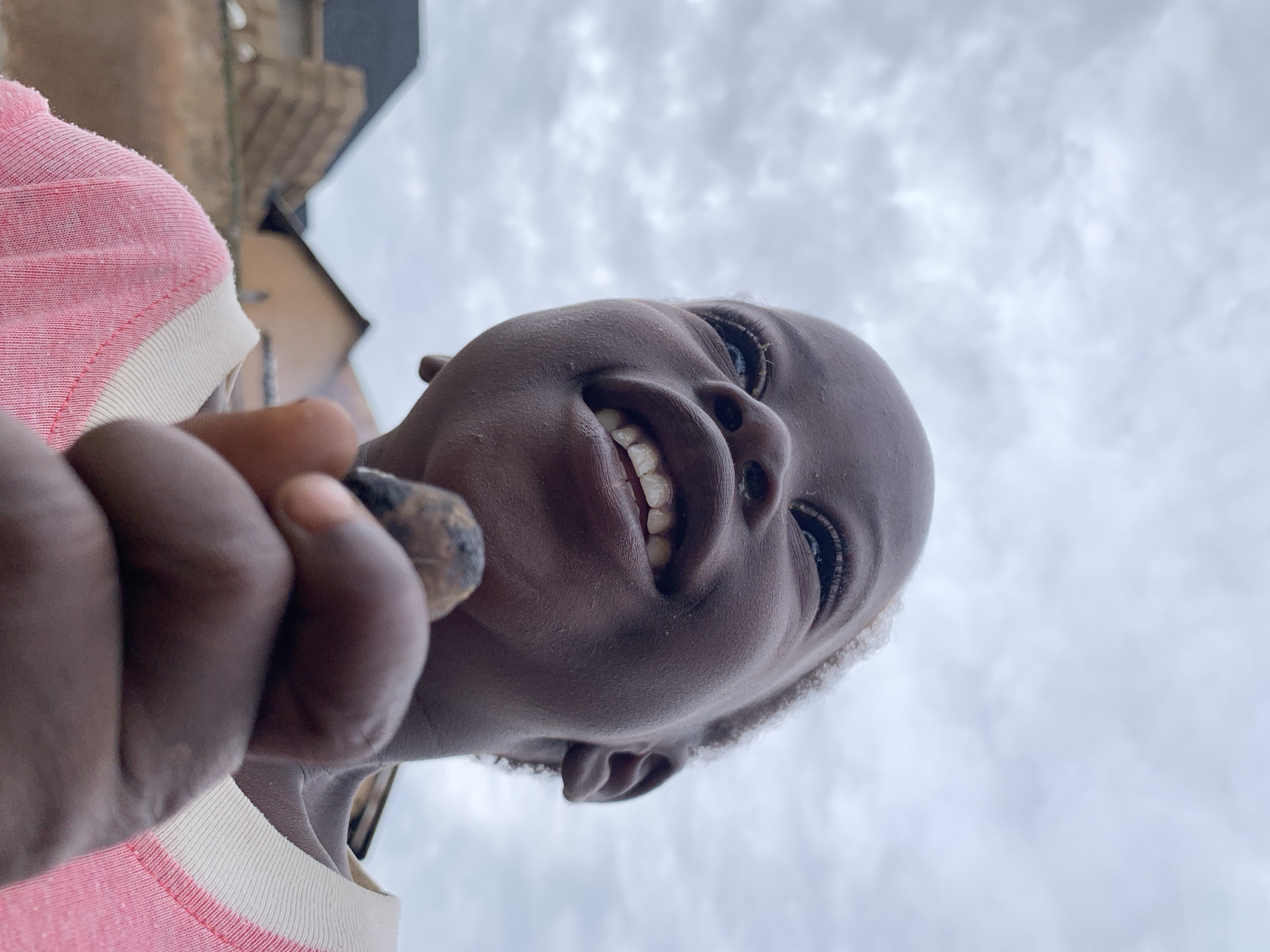A low-angle close-up shot captures a young Black child's face, looking upwards with a wide, joyful smile. The child's head is shaved, and they are wearing a pink t-shirt with white trim around the collar. In the foreground, the child's hand holds a small, dark object, possibly a seed or a piece of dirt, close to their mouth. In the background, out-of-focus buildings with brown and gray tones suggest an urban setting, consistent with the city of Jos, Nigeria. The sky above is overcast with soft, gray clouds.