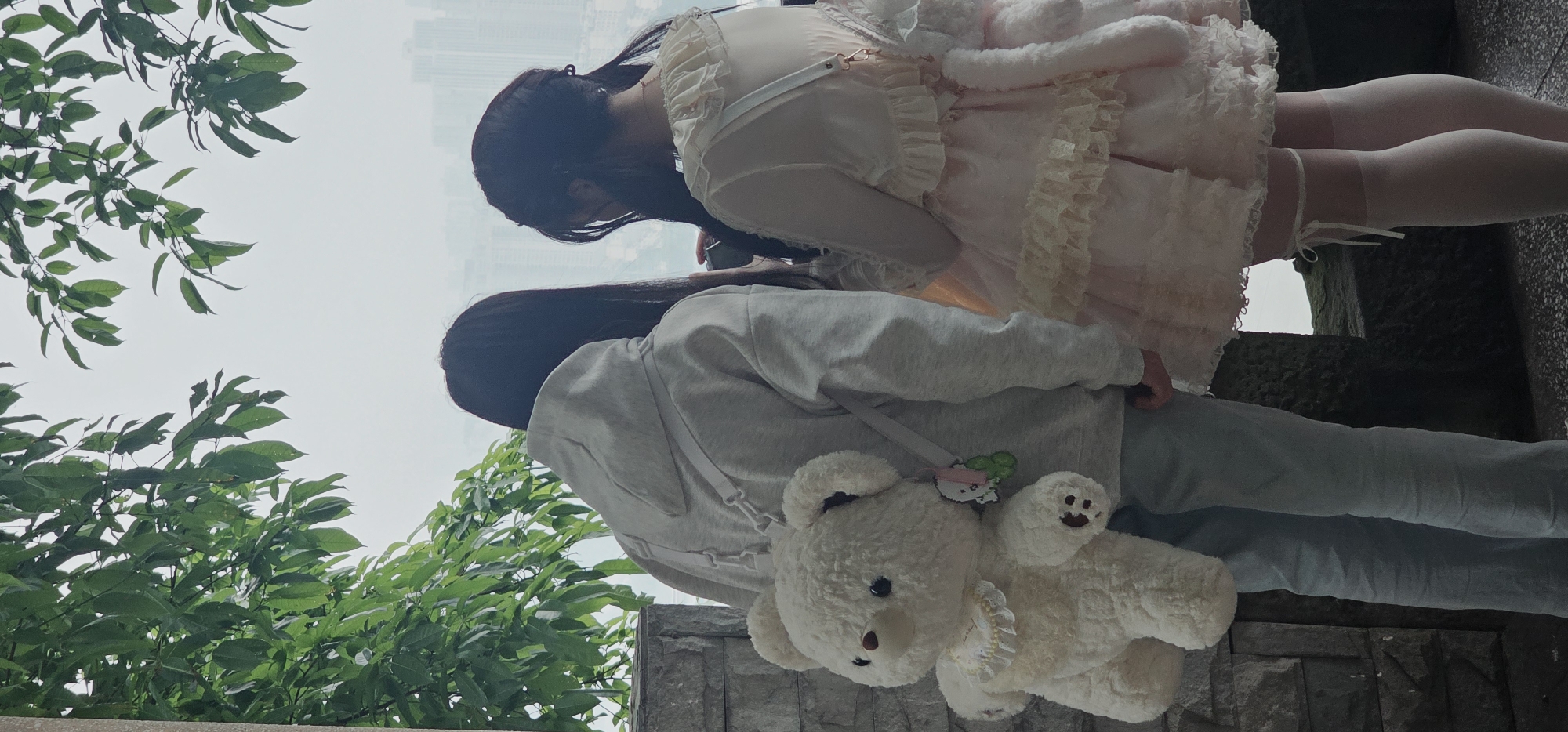 The image shows two young women in Yuzhong District, China. One woman is wearing a light pink, frilly dress with white tights and a garter belt. The other is wearing a grey hoodie and sweatpants, with a white teddy bear bag slung across her body. Both women have long, dark hair. Green foliage is visible on the left side of the image. In the background, indistinct city buildings are visible in the distance. The women are standing on a stone structure.