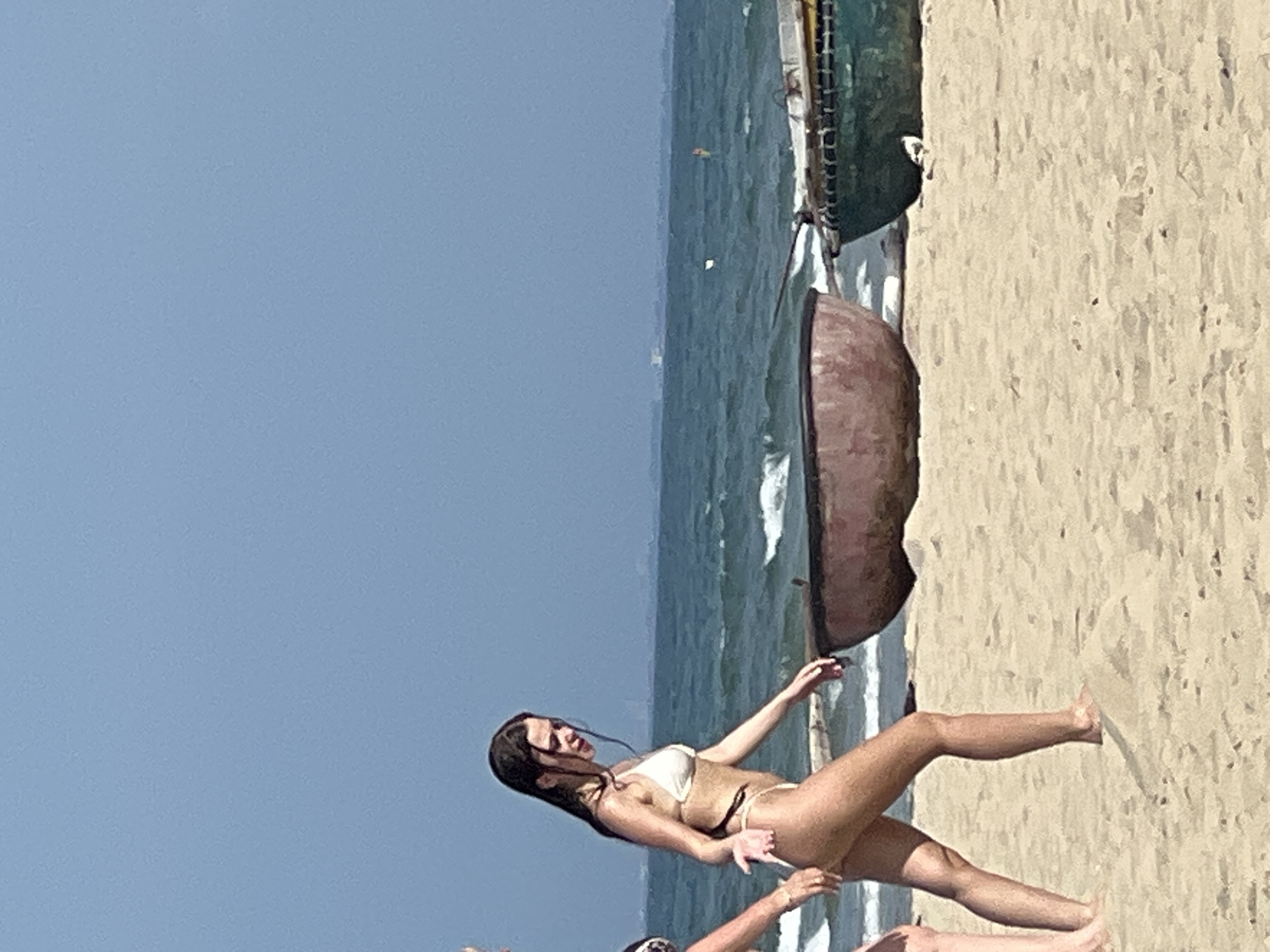 The media file shows a beach scene in Da Nang, Vietnam. The main subject is a young woman in a bikini standing on a sandy beach. She has long, dark hair and is looking to her left with a relaxed expression. Her arms are outstretched, with her left arm bent at the elbow and her right arm extended. She is wearing a light-colored bikini.
In the background, the ocean is visible with waves rolling in. A small, overturned boat, possibly a fishing boat, is situated on the beach near the water. The sky is a clear, bright blue, suggesting a sunny day. The sand is a light tan color, with some texture and small dark specks. Another person's legs are visible in the lower right corner, suggesting that the woman is not alone.
The lighting indicates it is daytime, likely midday or afternoon due to the bright sun and shadows. The overall mood of the image is serene and peaceful, typical of a beach vacation. There is no visible text in the image. The presence of the overturned boat might suggest a working fishing community or a historical element of the location.